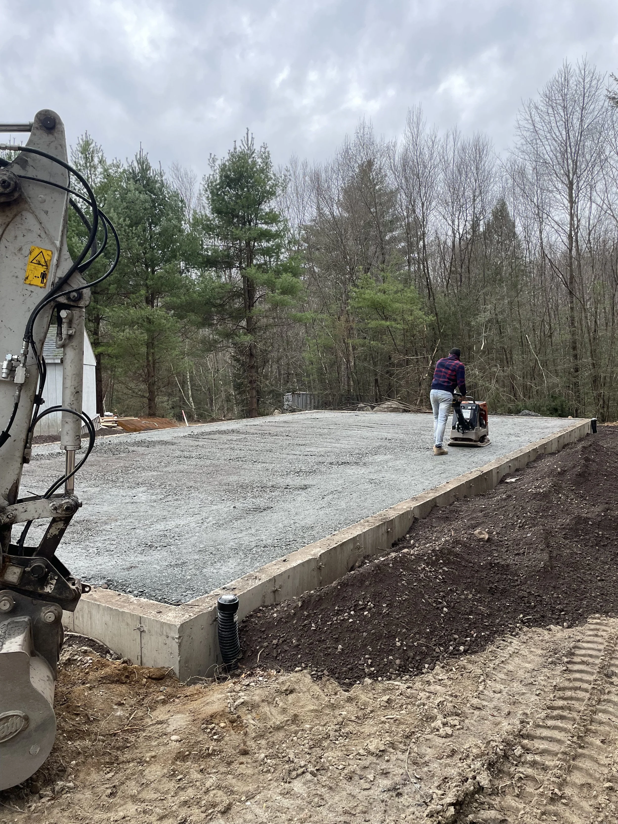 A construction worker operates a compactor machine on freshly laid gravel on a construction site outdoors, with a wooded background and cloudy sky overhead.