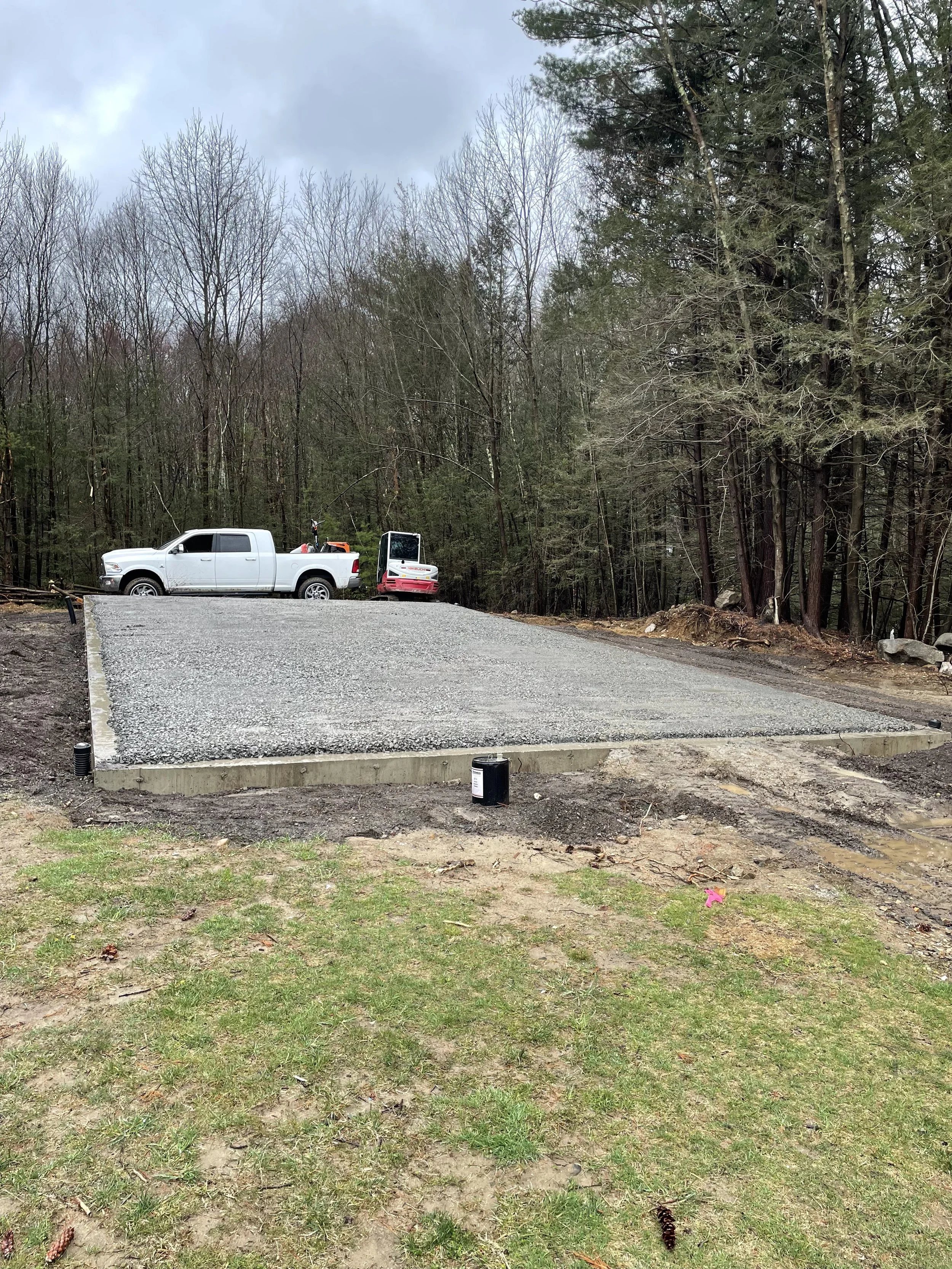 Construction site with a gravel foundation, a white truck, and construction equipment, surrounded by wooded area with trees.
