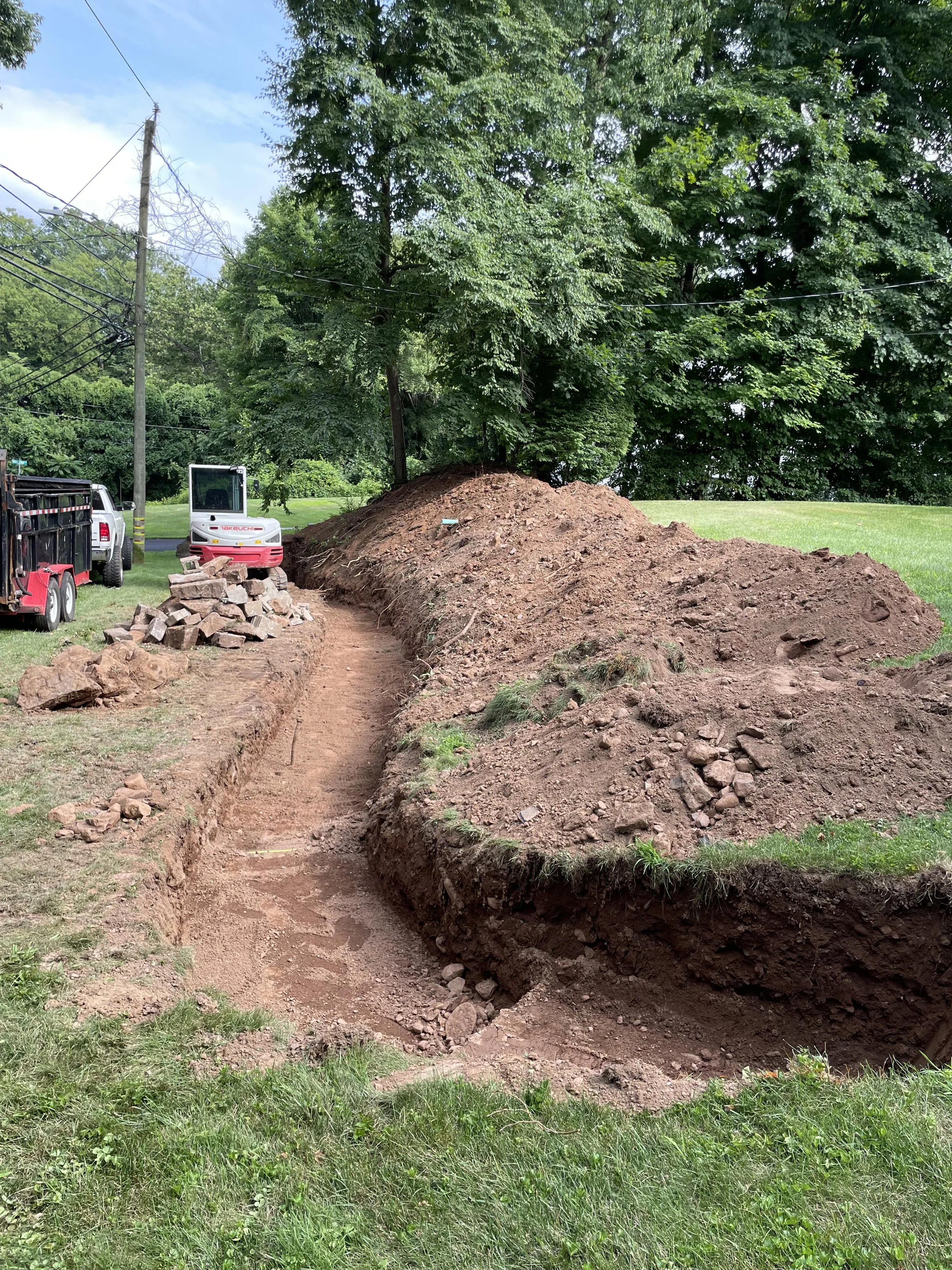 A construction site with a cleared dirt pathway next to large piles of dirt and rocks, nearby utility poles, trees, and construction equipment such as a small excavator and trucks.