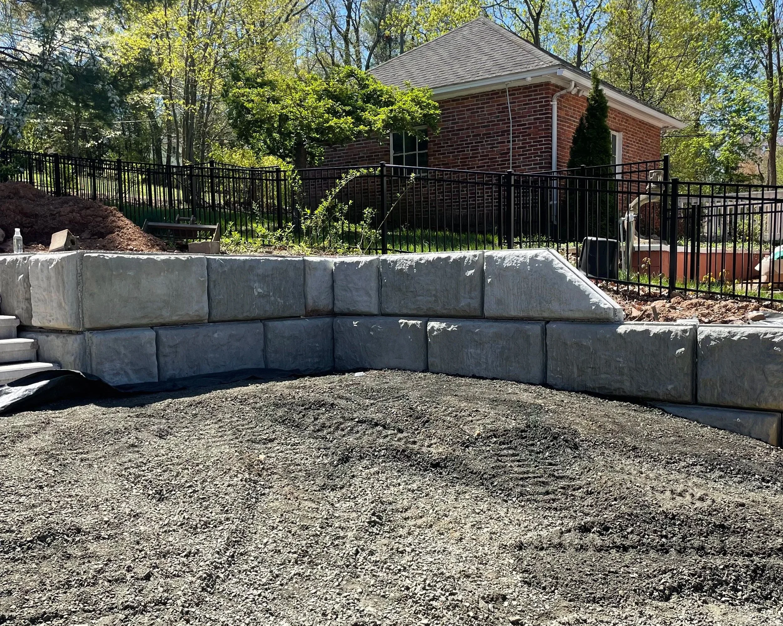 Construction site with a new stone retaining wall and stairs, a gravel pathway in the foreground, a fenced yard with trees, and a brick house in the background.