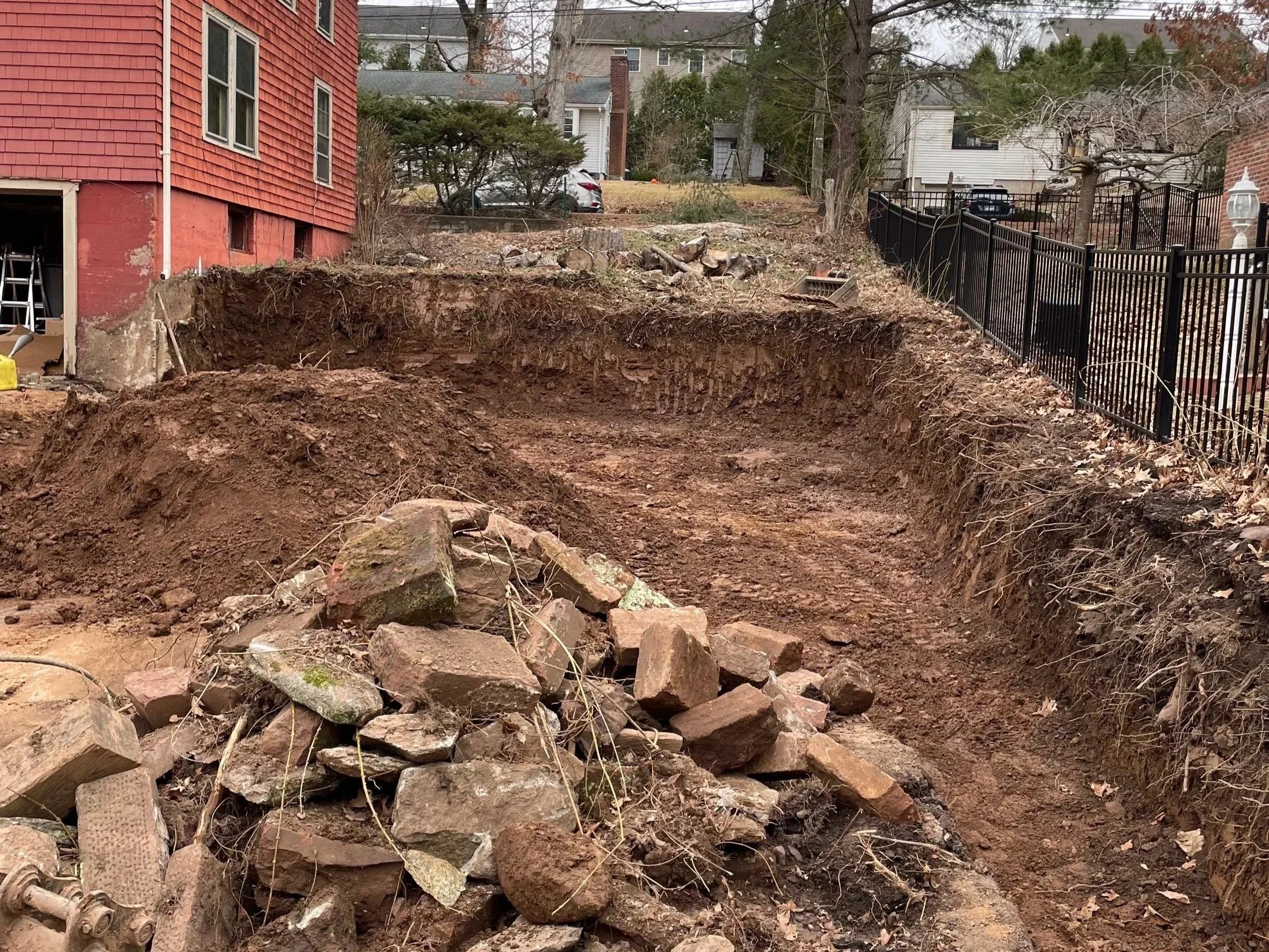 A backyard under construction with a large dugout area, piles of dirt and rocks, and a black metal fence along the yard's edge.
