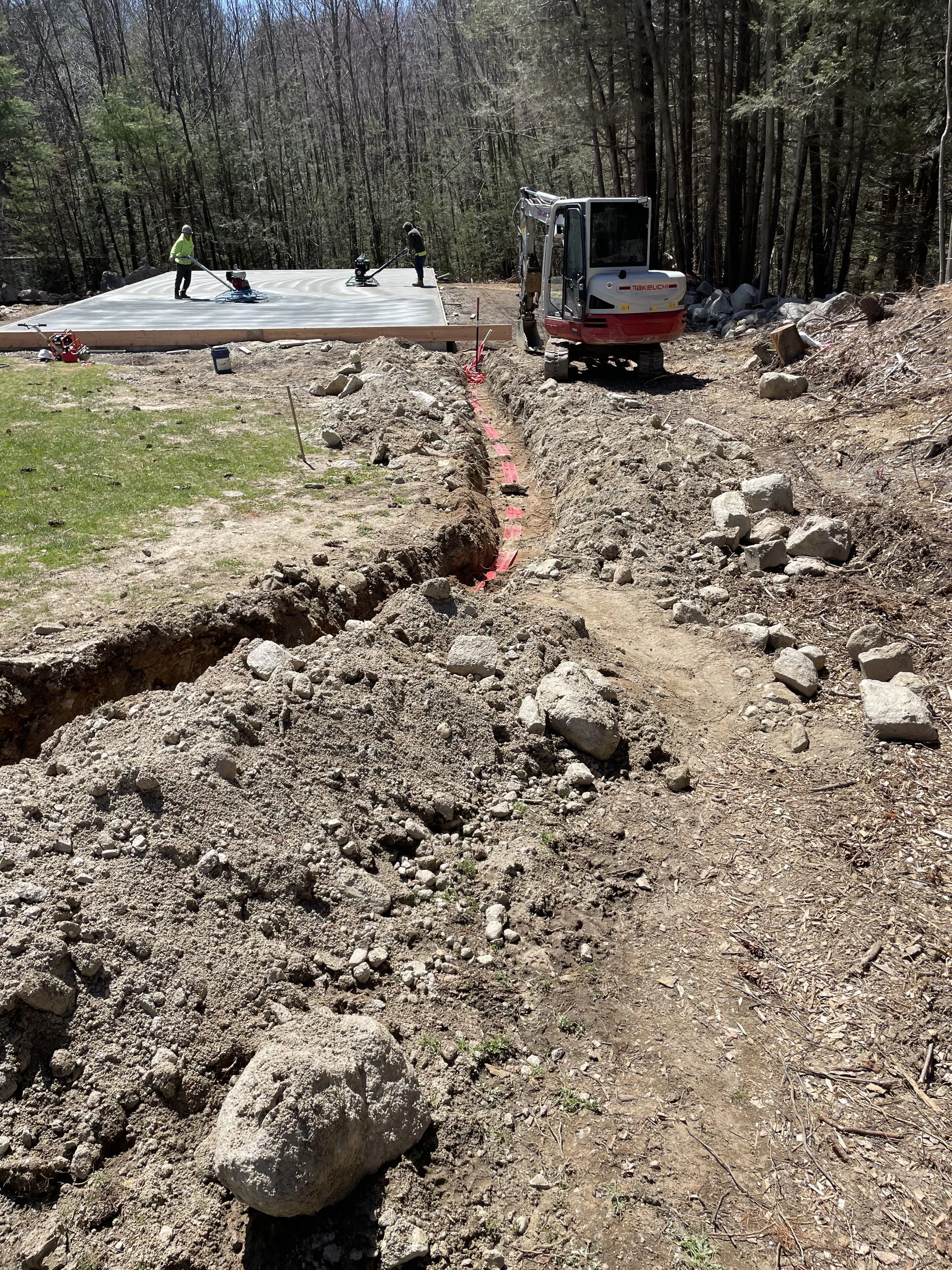Construction workers and a small excavator are working on a construction site in a wooded area, with a trench dug alongside a dirt path, and a section of the ground prepared for laying a foundation or pavement.