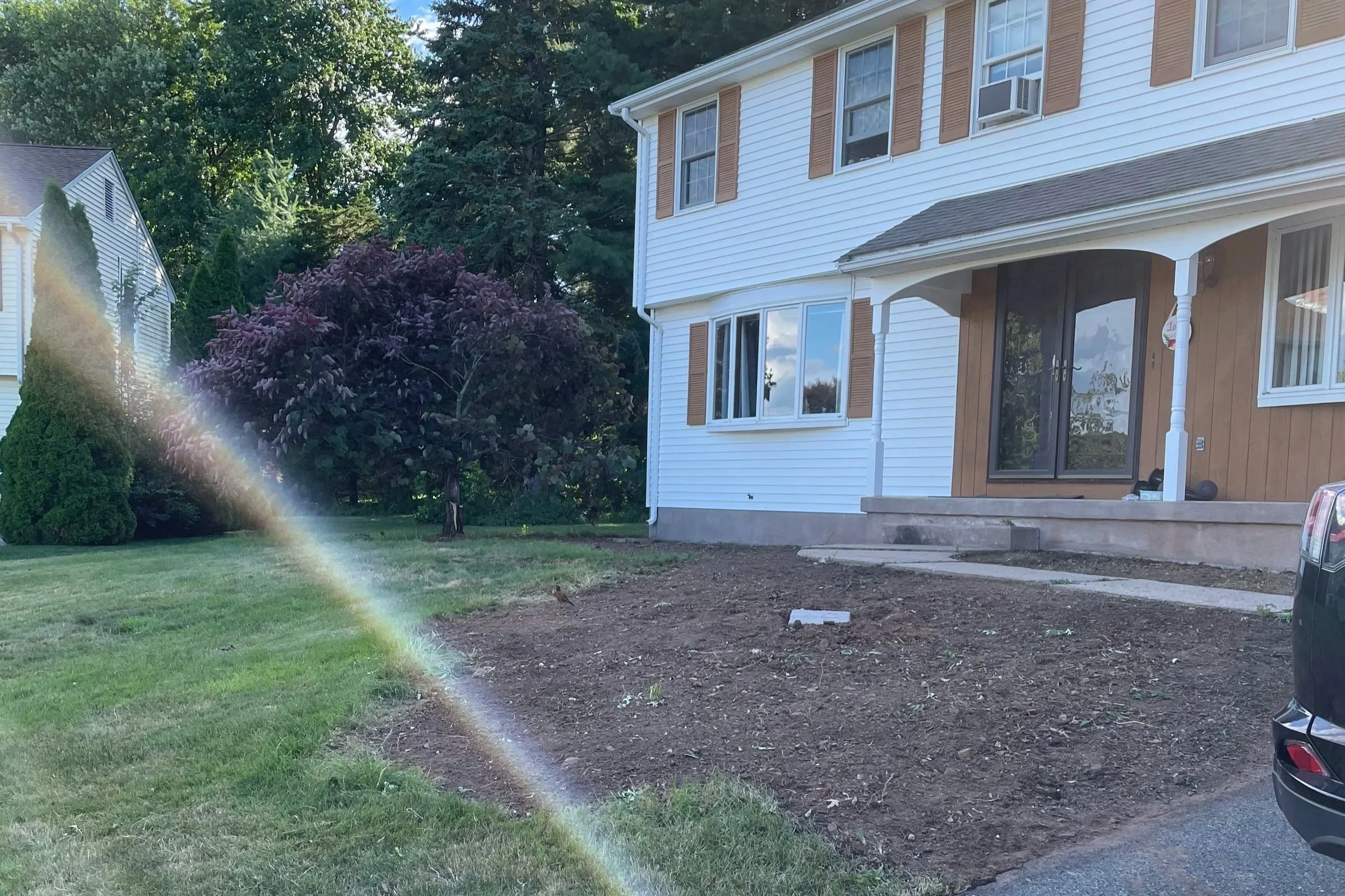 Front yard of a house with a patch of bare soil, a concrete pathway, and a driveway with a car. The house has white siding, brown shutters, and a small porch with steps.
