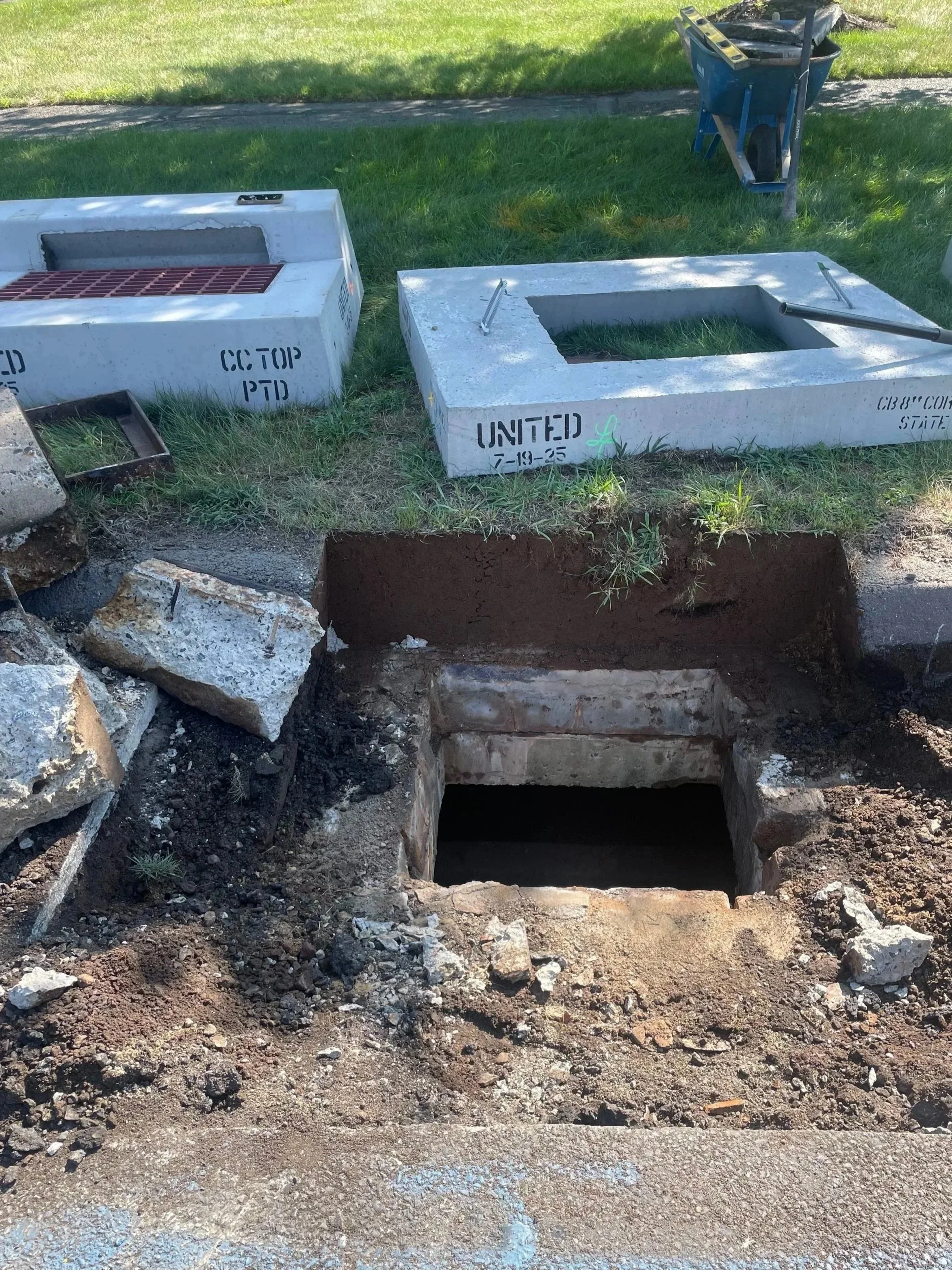 Open manhole or utility access hole in the ground with construction bricks and dirt around it, and tools and covers placed nearby on the grass.