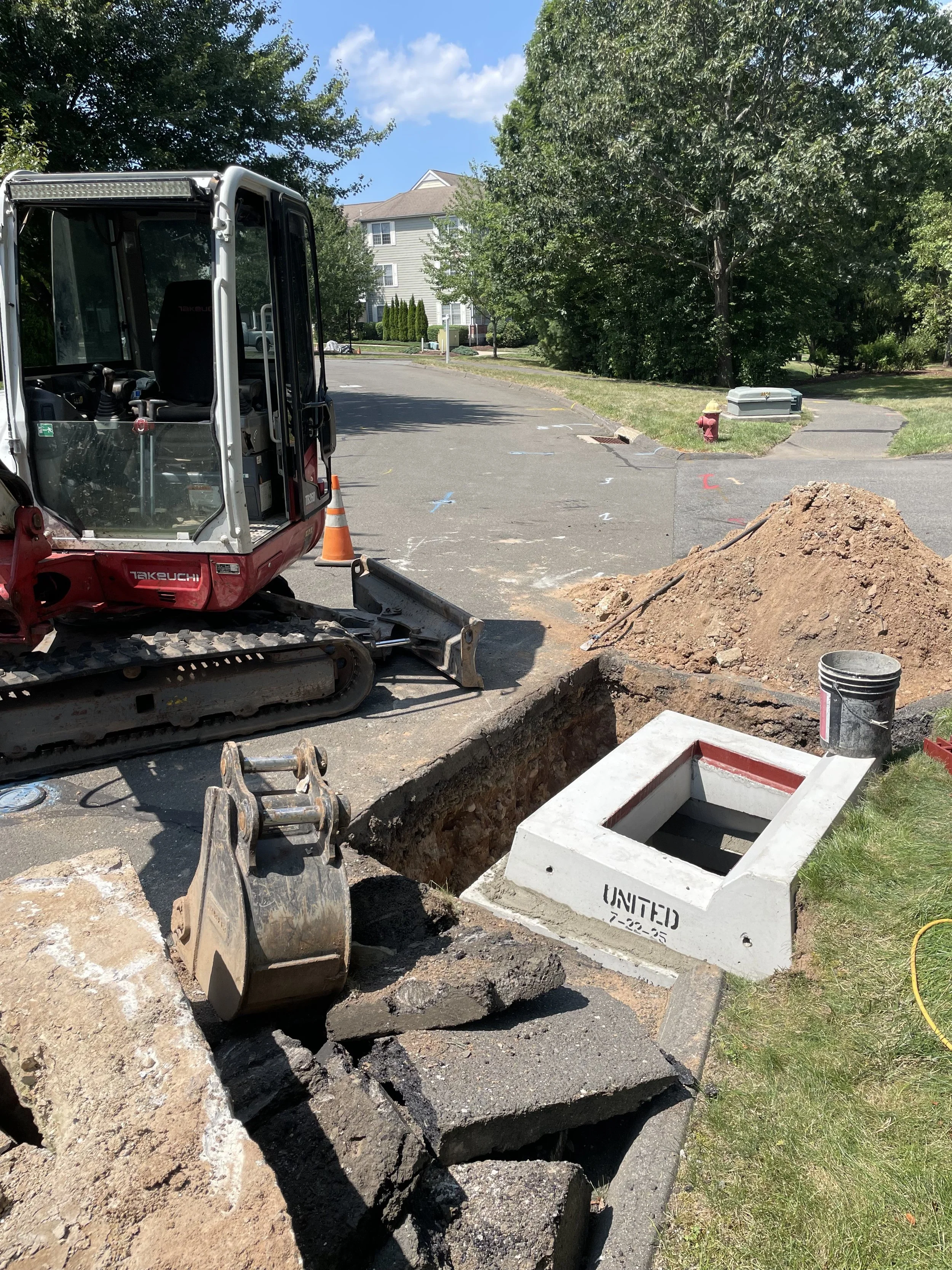 Construction site with an excavator digging a trench, orange cone nearby, and a manhole with a concrete cover labeled 'UNITED' on a residential street with trees and houses.