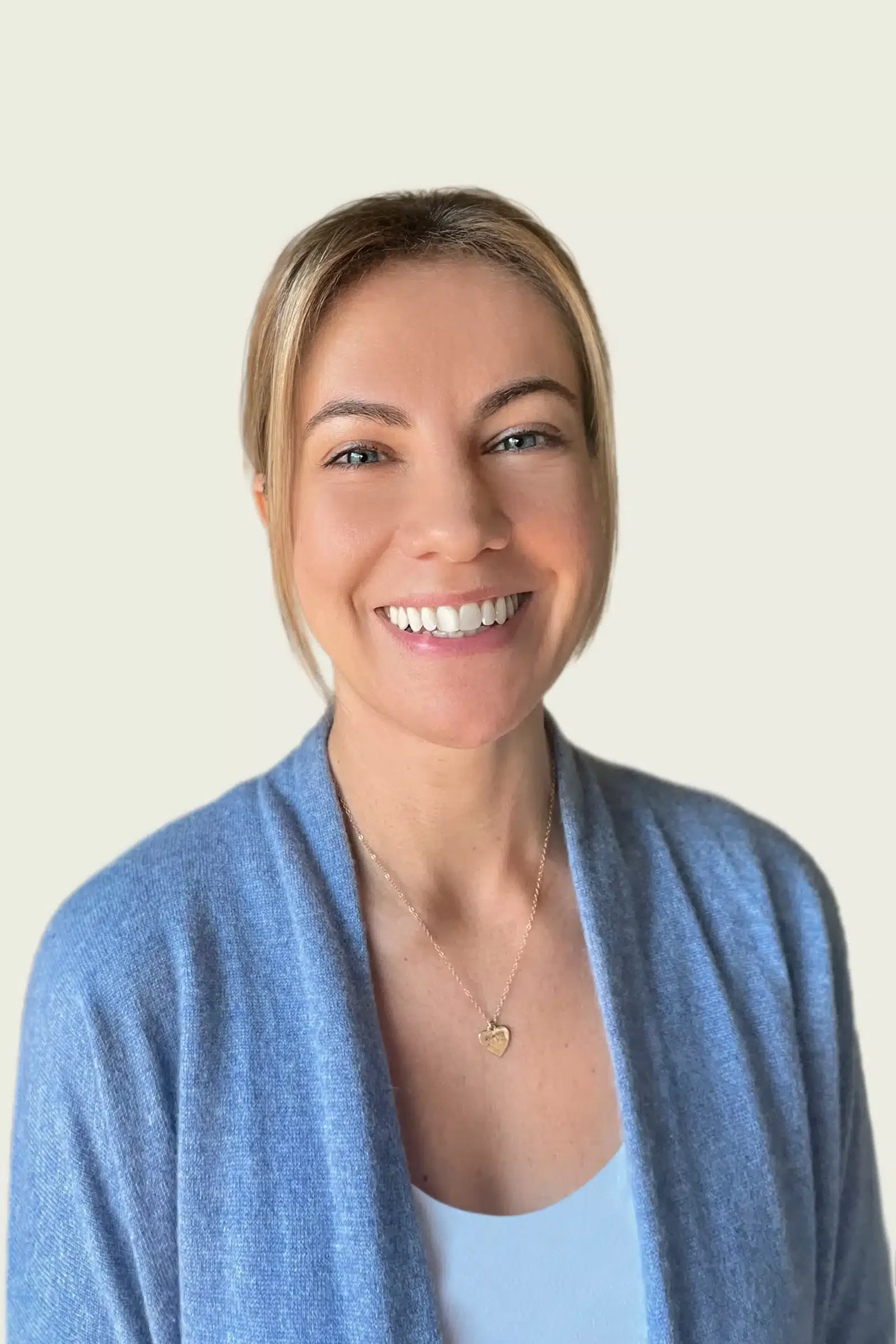 A woman with short blonde hair smiling, wearing a blue cardigan and a white top, with a gold heart necklace, standing against a light-colored background.