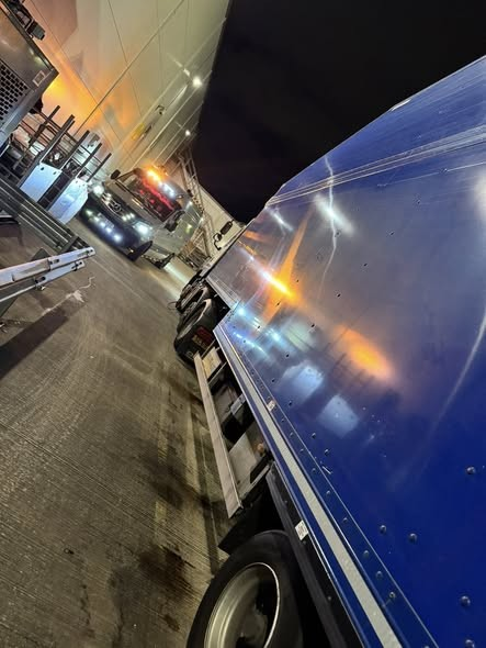 A large blue semi-truck parked at a night loading dock, with other trucks and equipment visible in the background.
