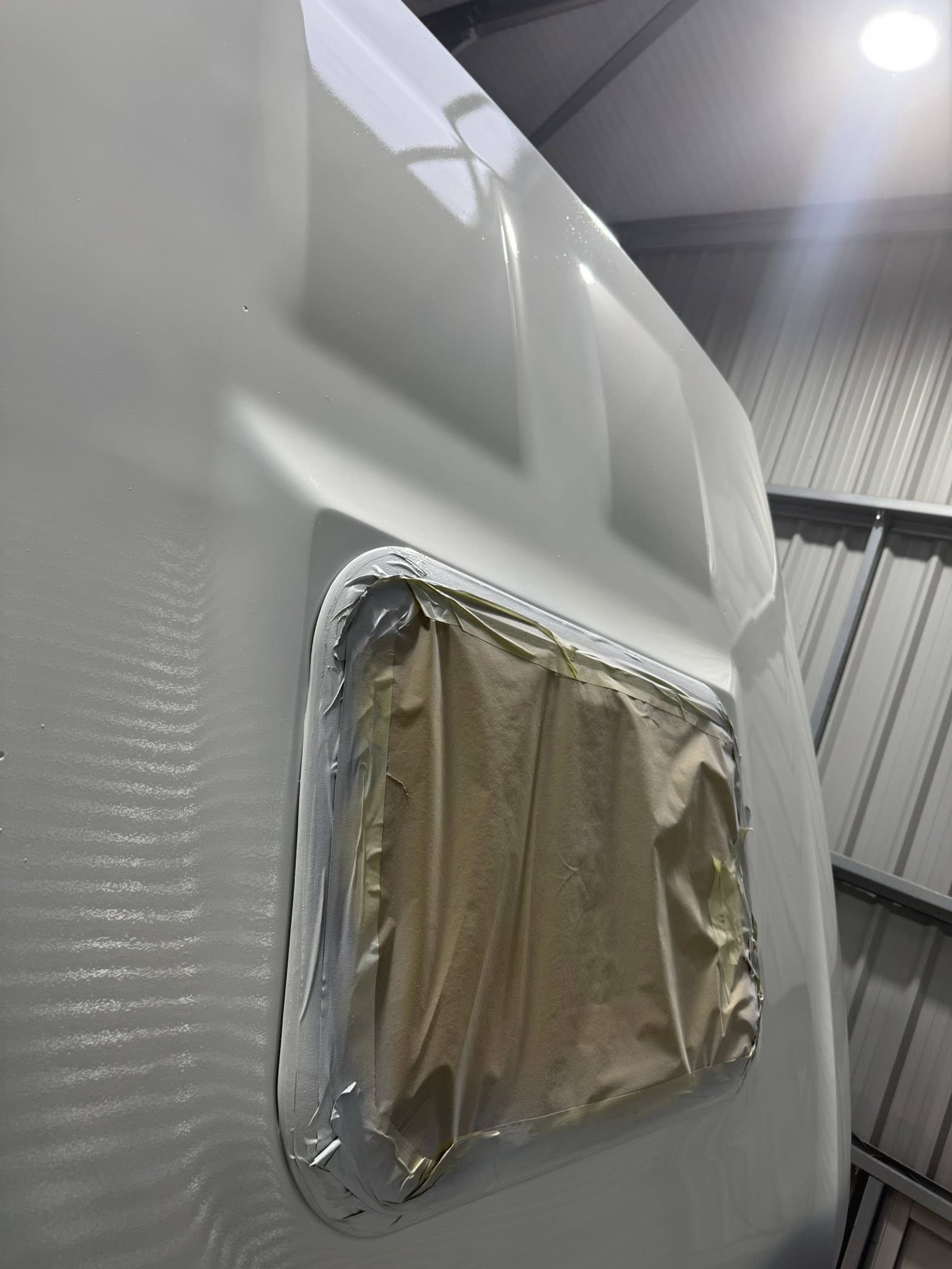 A close-up of a white vehicle with a covered opening, possibly a window or panel, taped off with painter's tape and paper, in a warehouse or garage setting.