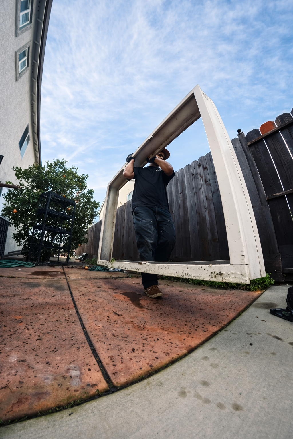 A person in black clothing lifting a large plastic frame over their shoulders in a backyard with a wooden fence and house nearby, under a partly cloudy sky.