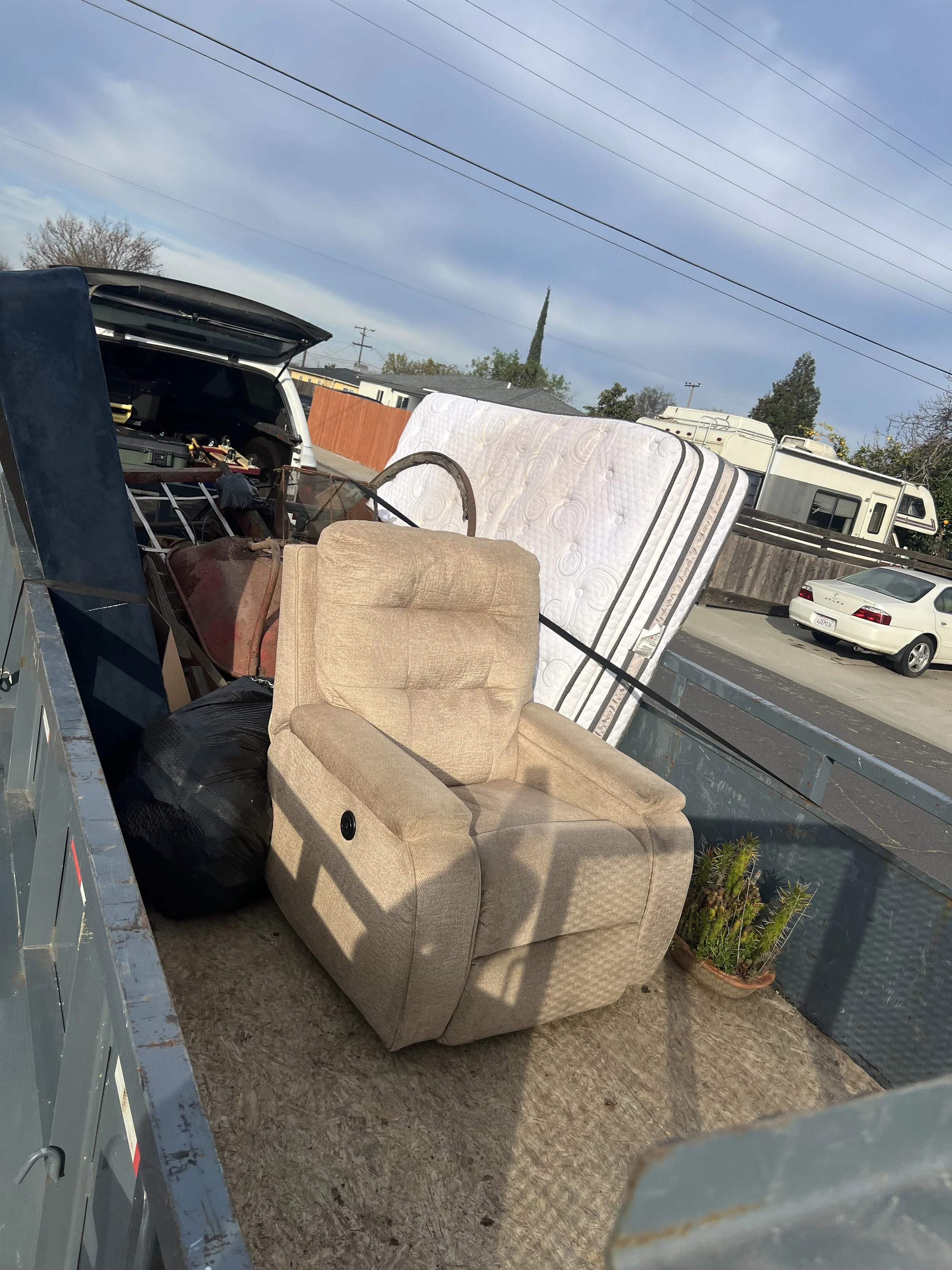 A beige armchair, a mattress, a black trash bag, and other items loaded in a truck bed, with residential houses and a street in the background.