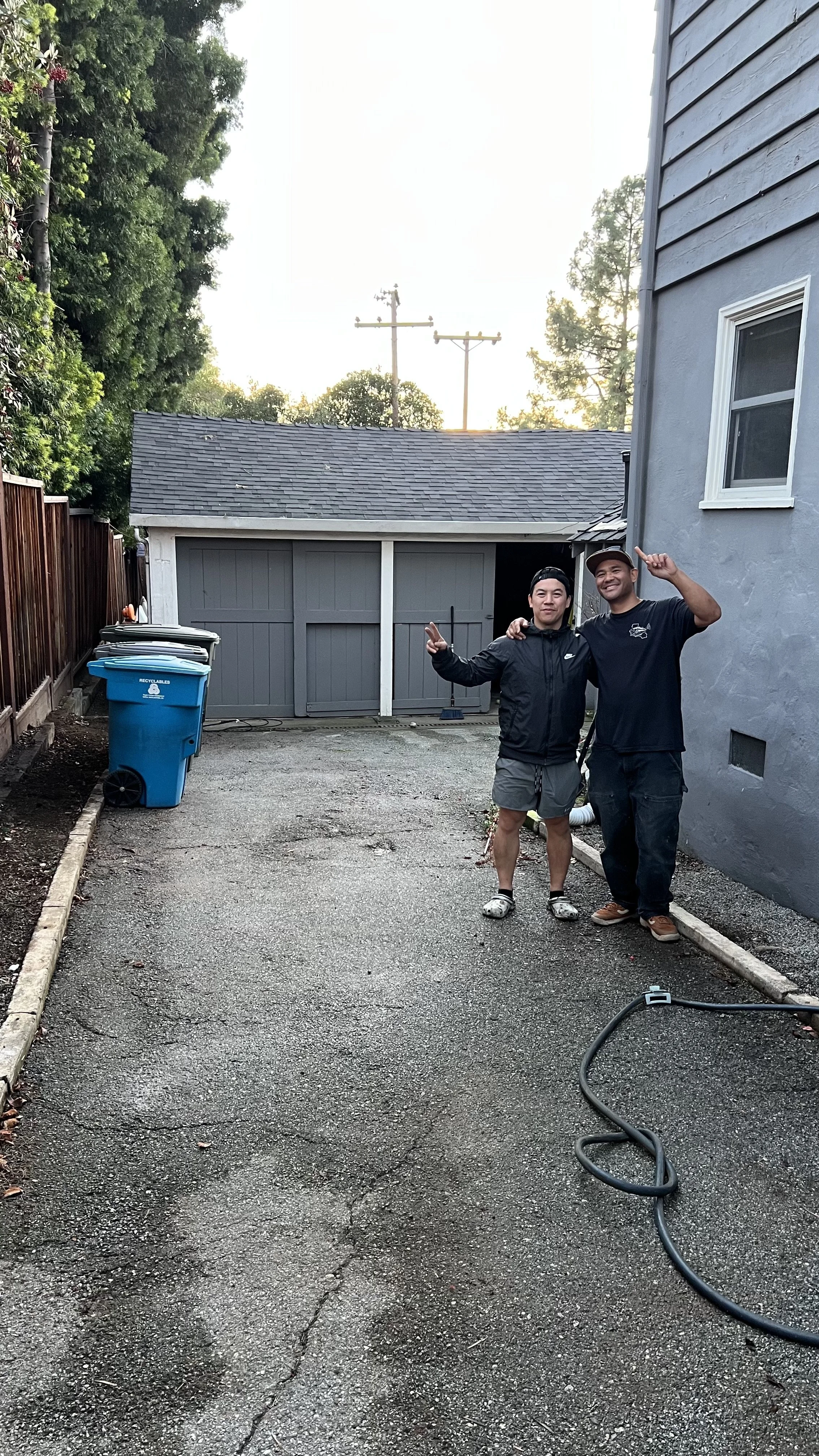 Two men standing on a driveway, smiling and making peace signs, with a gray garage and trees in the background.