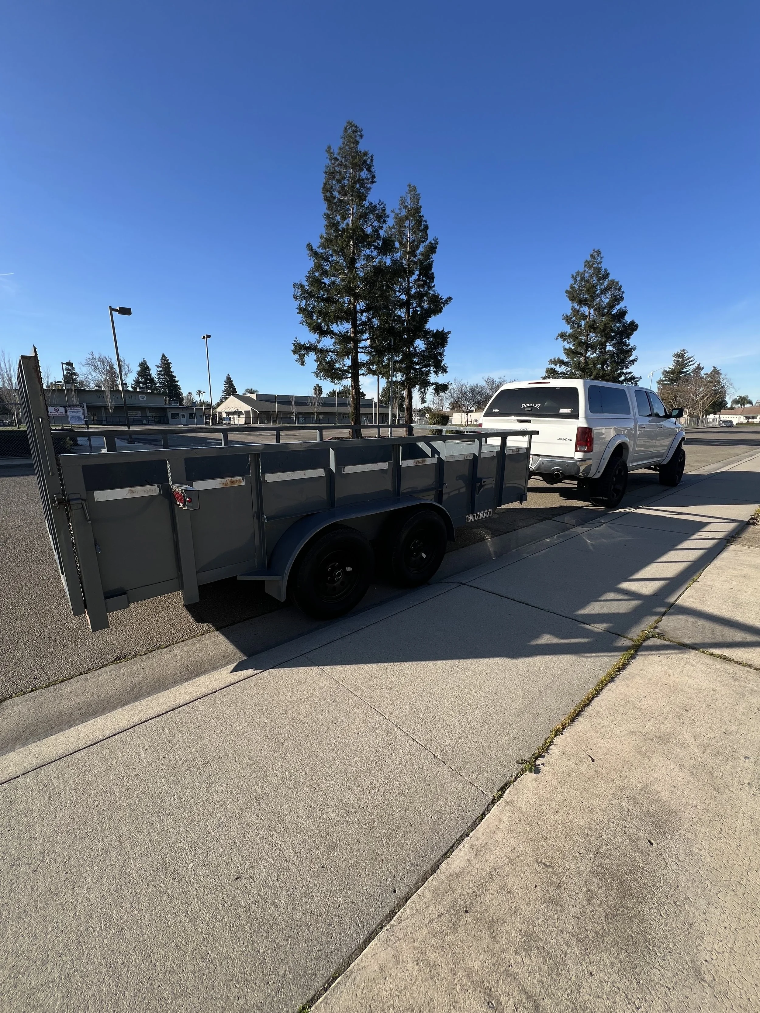 White pickup truck attached to a flatbed trailer parked on a sidewalk in a suburban area with a clear blue sky and tall trees in the background.