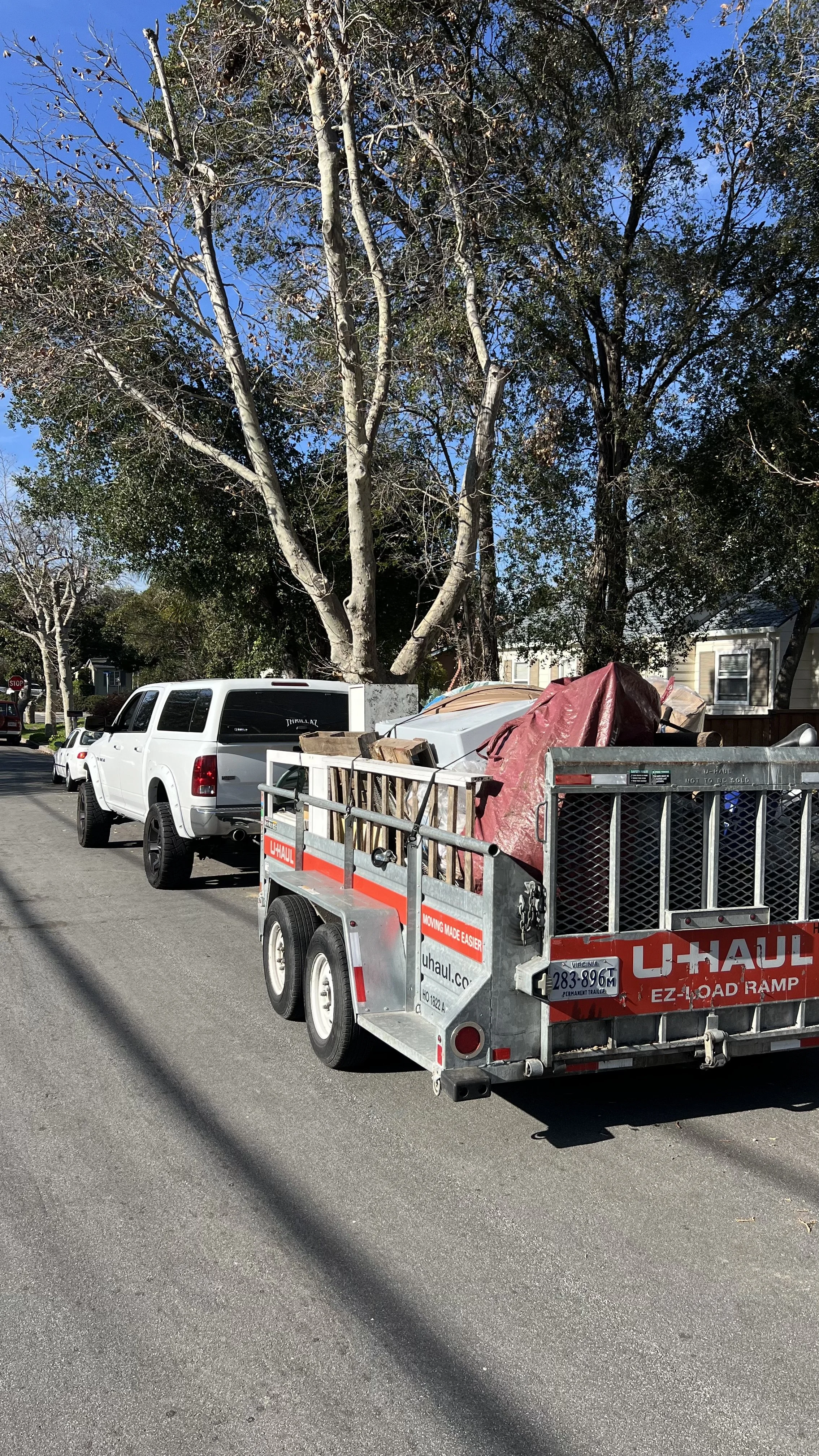 A U-Haul trailer attached to a white pickup truck parked on a residential street, with a large tree and houses in the background. The trailer is loaded with various items, including a red tarp, wood, and other belongings.