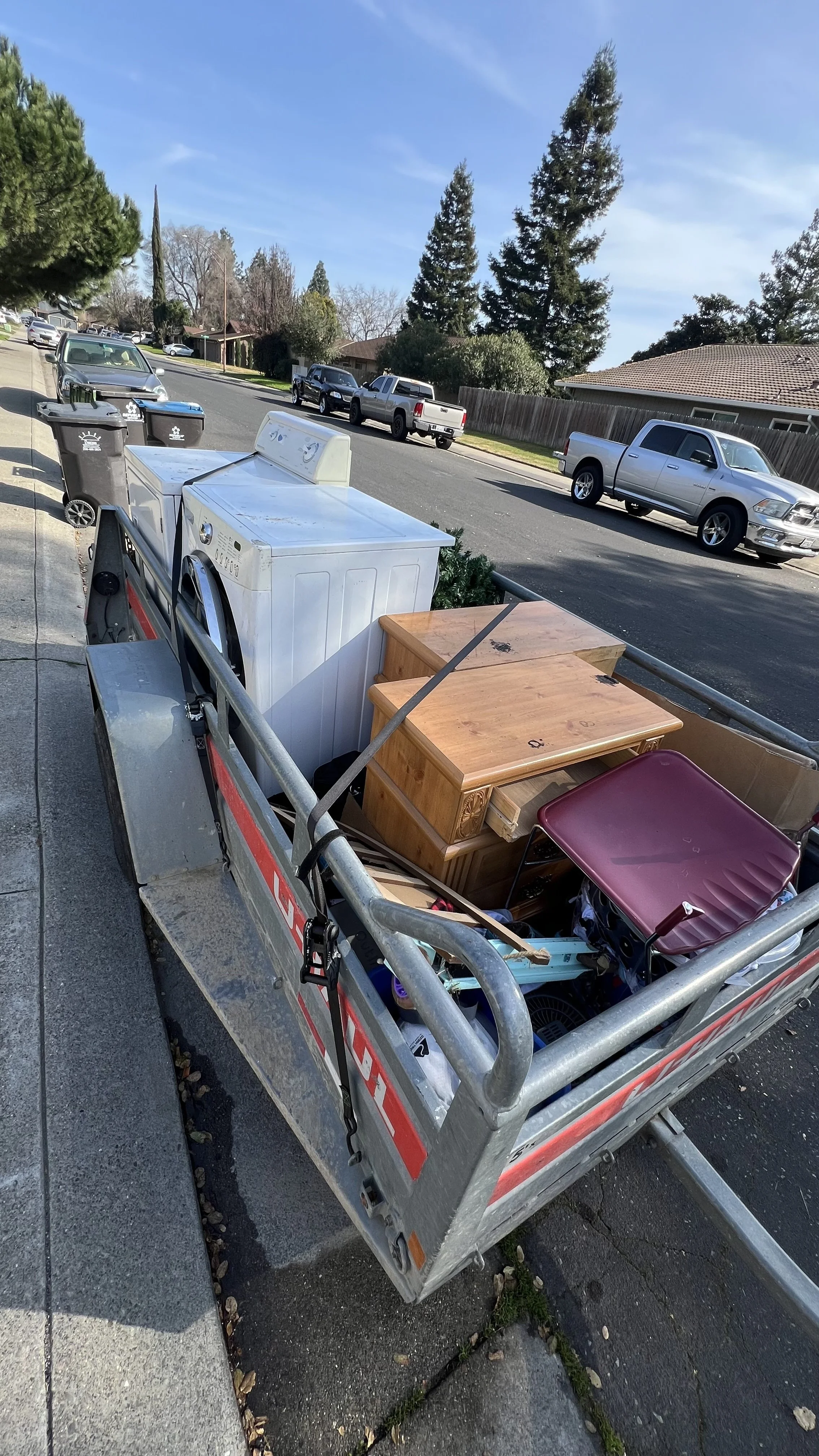 A utility trailer parked on a sidewalk, loaded with a washing machine, wooden furniture, and various household items, with a residential street and parked cars in the background.