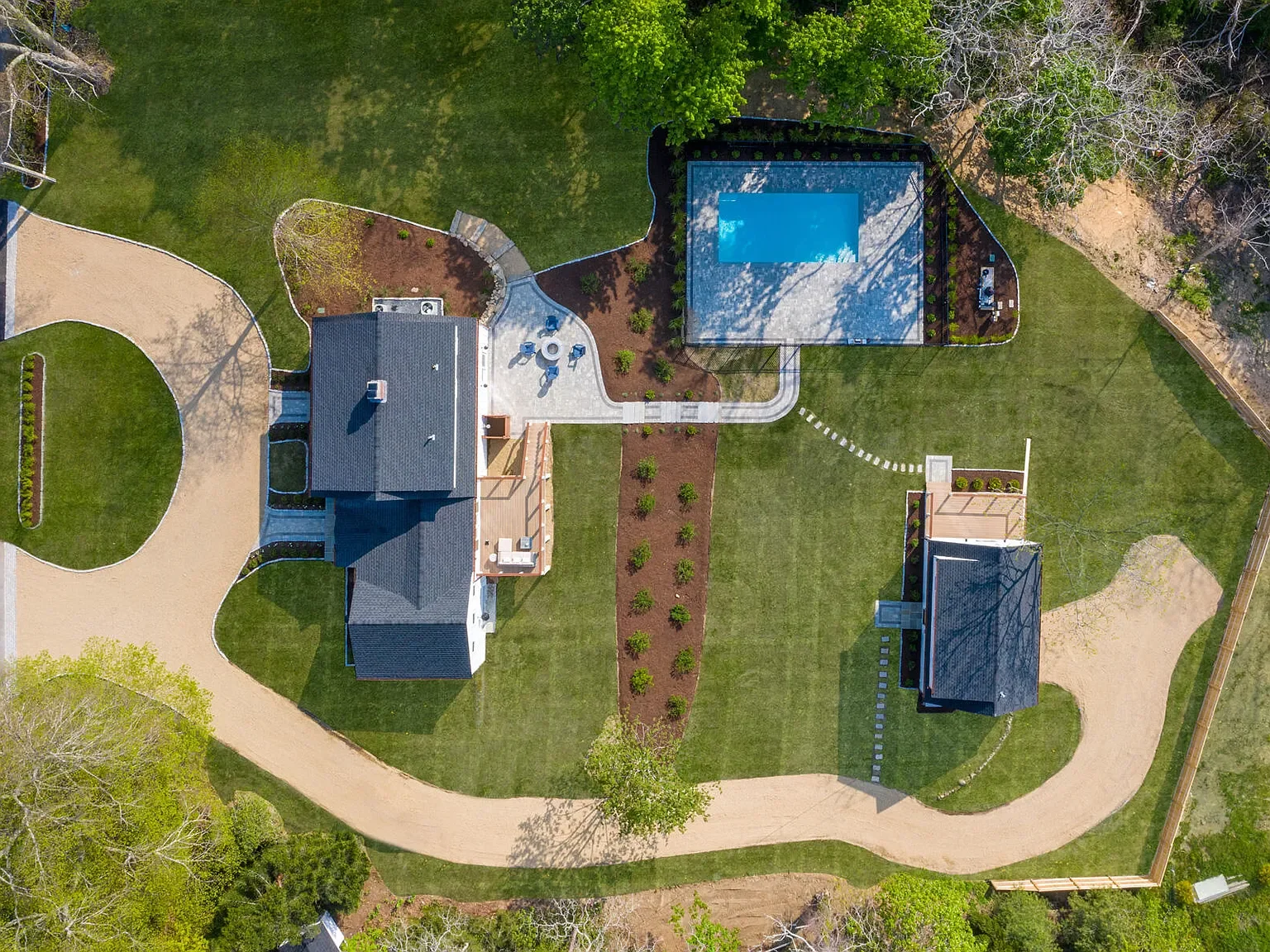 Aerial view of a backyard showing two houses, a swimming pool, a lawn, a paved patio with outdoor furniture, a dirt driveway, and surrounding trees.