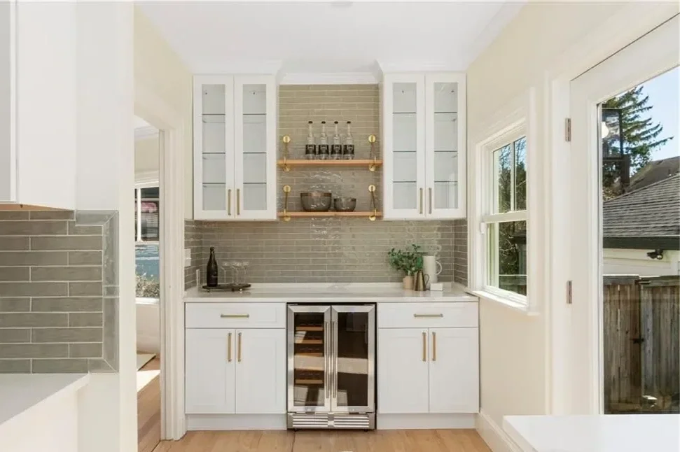 Kitchen with white cabinets, glass-front upper cabinets, and gray brick backsplash. Contains a small wine fridge, a bottle, a plant, and some bowls. Large window shows outdoor trees and a neighboring house.