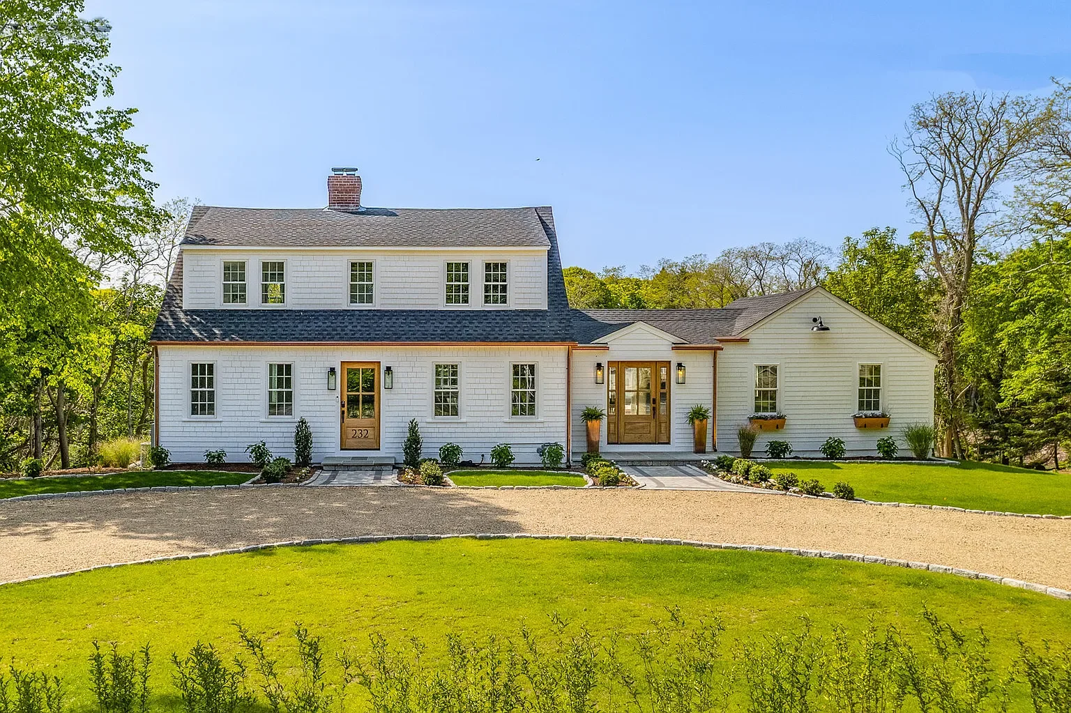 White farmhouse-style house with landscaped front yard, paved driveway, and surrounded by trees.