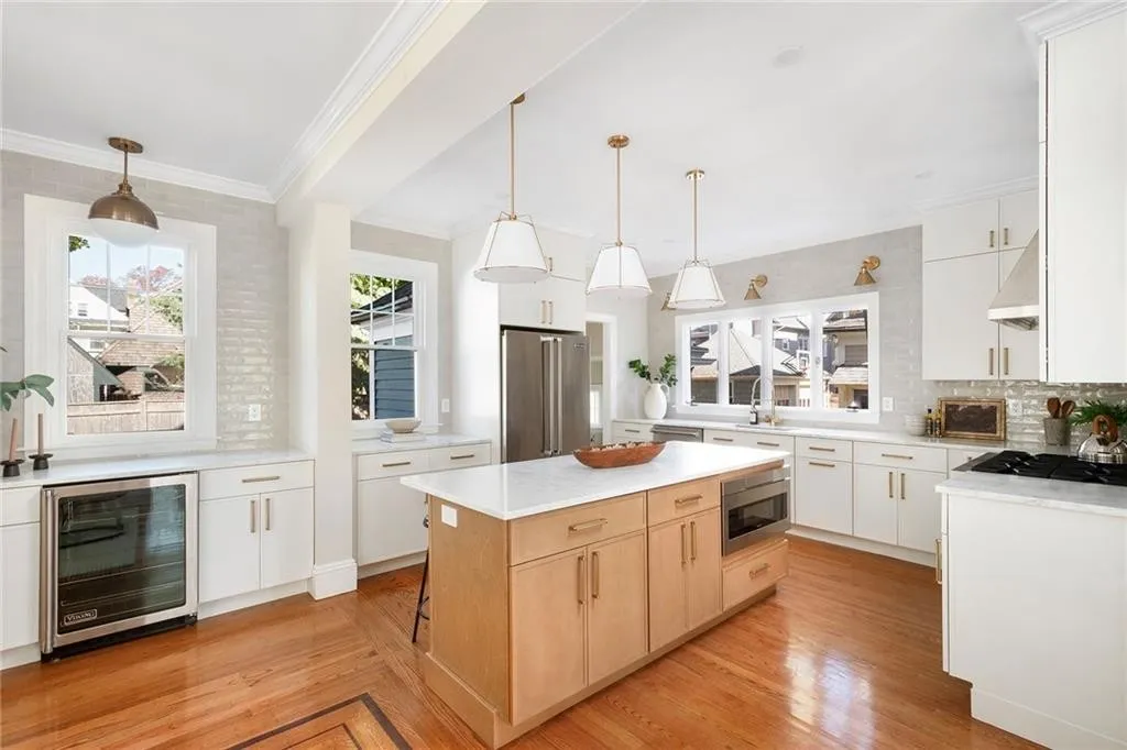 Bright, modern kitchen with white cabinets, wooden flooring, central island, stainless steel appliances, and large windows letting in natural light.