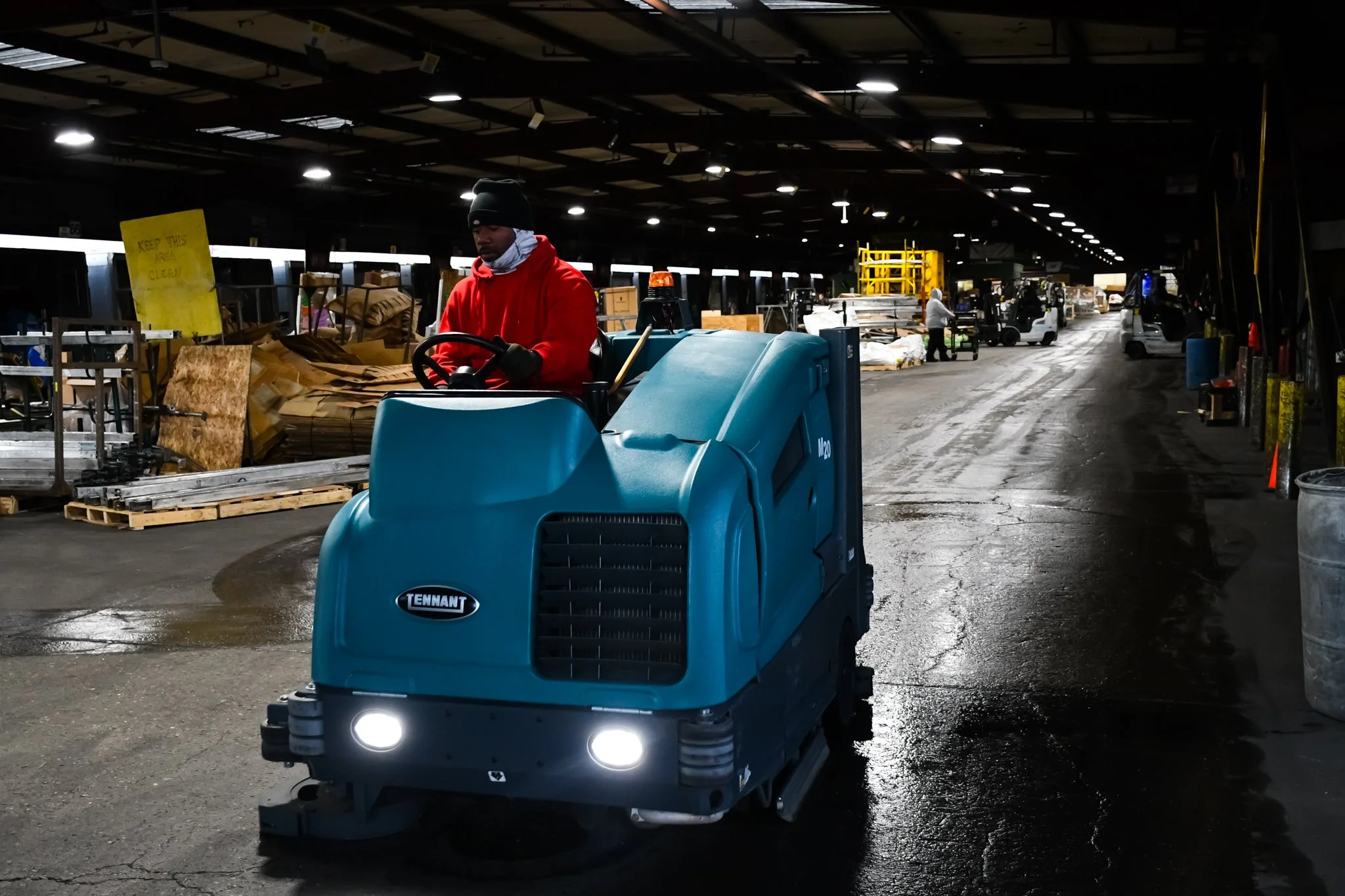 Man on commercial floor scrubbing machine cleans warehouse floor leaving trails of wet, clean concrete.