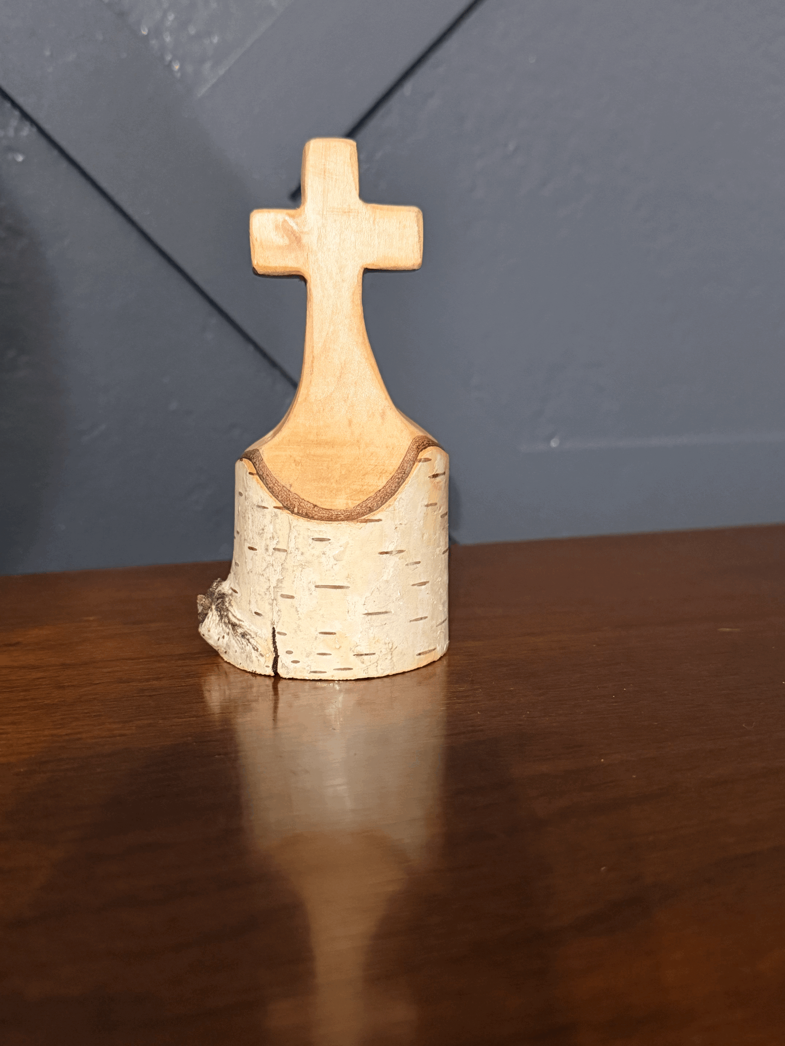 Small handcrafted wooden cross standing in a natural wood base with bark detail, measuring approximately 2.5 by 4 inches.
