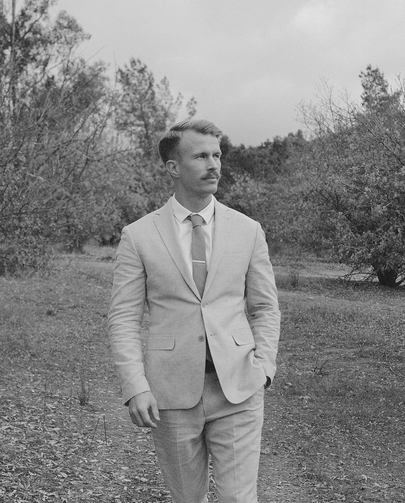 A man in a light-colored suit with a tie, standing outdoors in a natural landscape with trees and a dirt path.