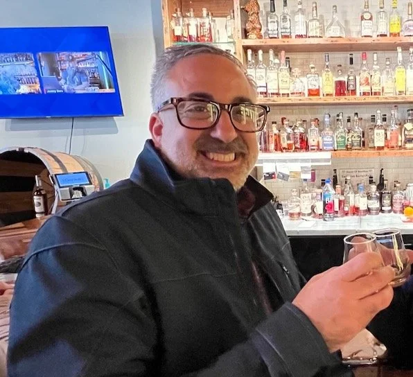 A man with glasses smiling and holding a glass of whiskey in a bar with shelves of liquor bottles behind him.