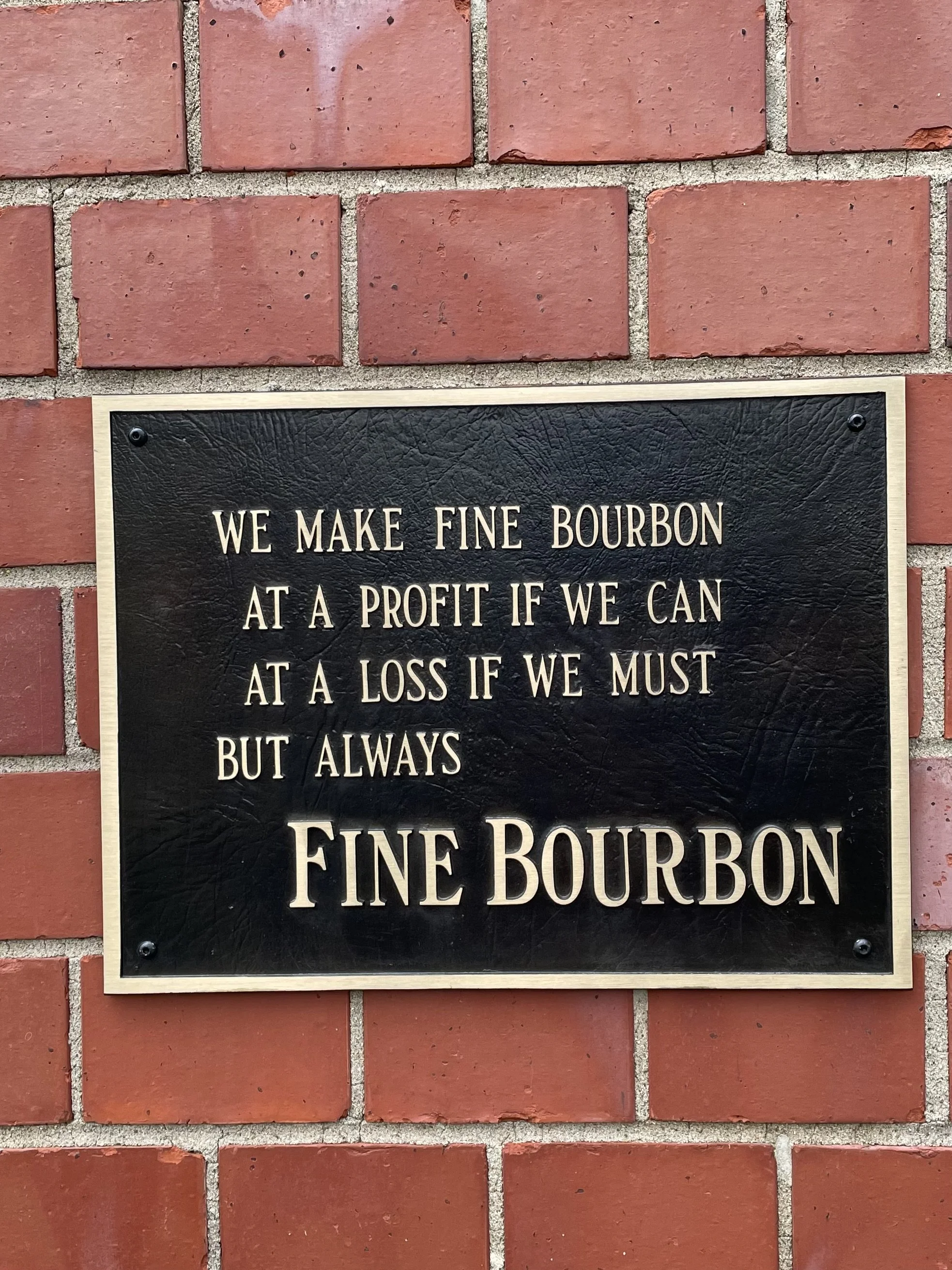 A black sign with beige text mounted on a brick wall. The sign reads: 'We make fine bourbon at a profit if we can at a loss if we must but always fine bourbon.'