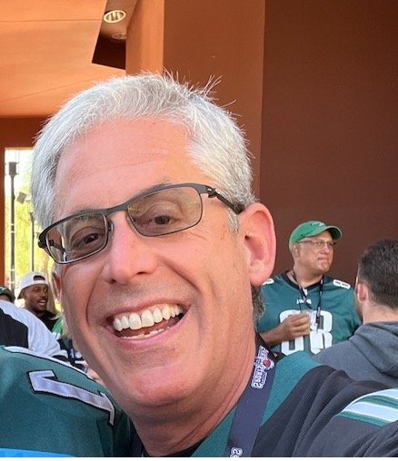 A man with gray hair and glasses smiling at a social gathering, wearing a sports jersey.