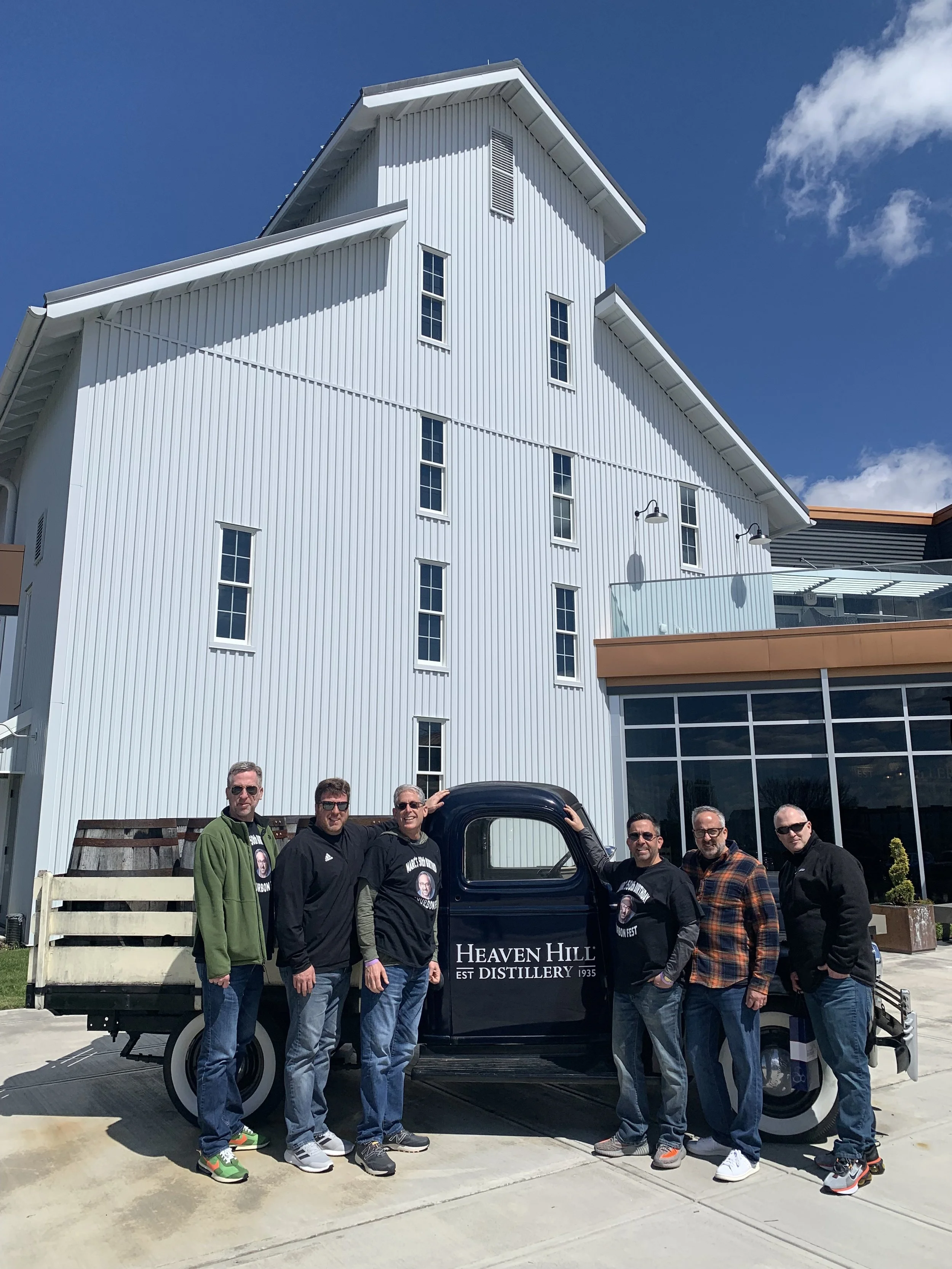 Seven men standing outdoors next to a vintage truck with 'Heaven Hill Est Distillery 1935' written on it, in front of a large white building under a blue sky.