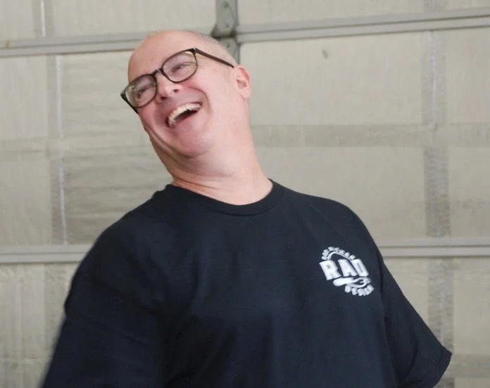A man with glasses laughing and smiling, wearing a black t-shirt, in front of a garage door.