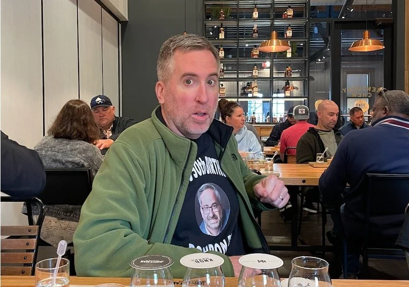 A man with short gray hair and a beard, wearing a green jacket over a graphic t-shirt, sitting at a wooden table in a busy restaurant or cafe. The background features other diners, shelving with bottles, and modern decor.