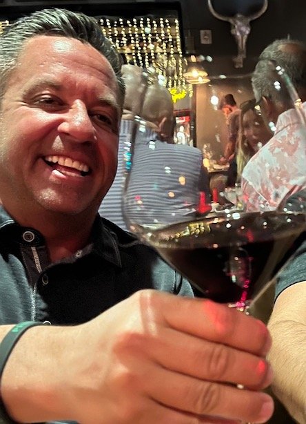 Person smiling and holding a glass of red wine in a lively restaurant or bar with people in the background.