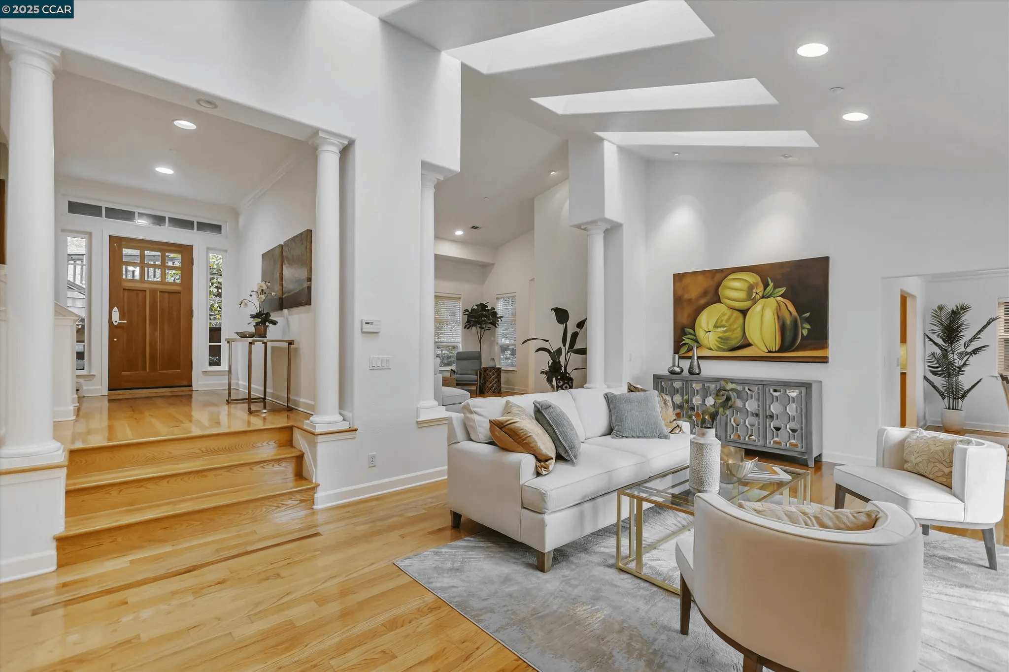 Living room with white walls, hardwood floors, modern furniture, a painting of green fruit, and decorative plants.