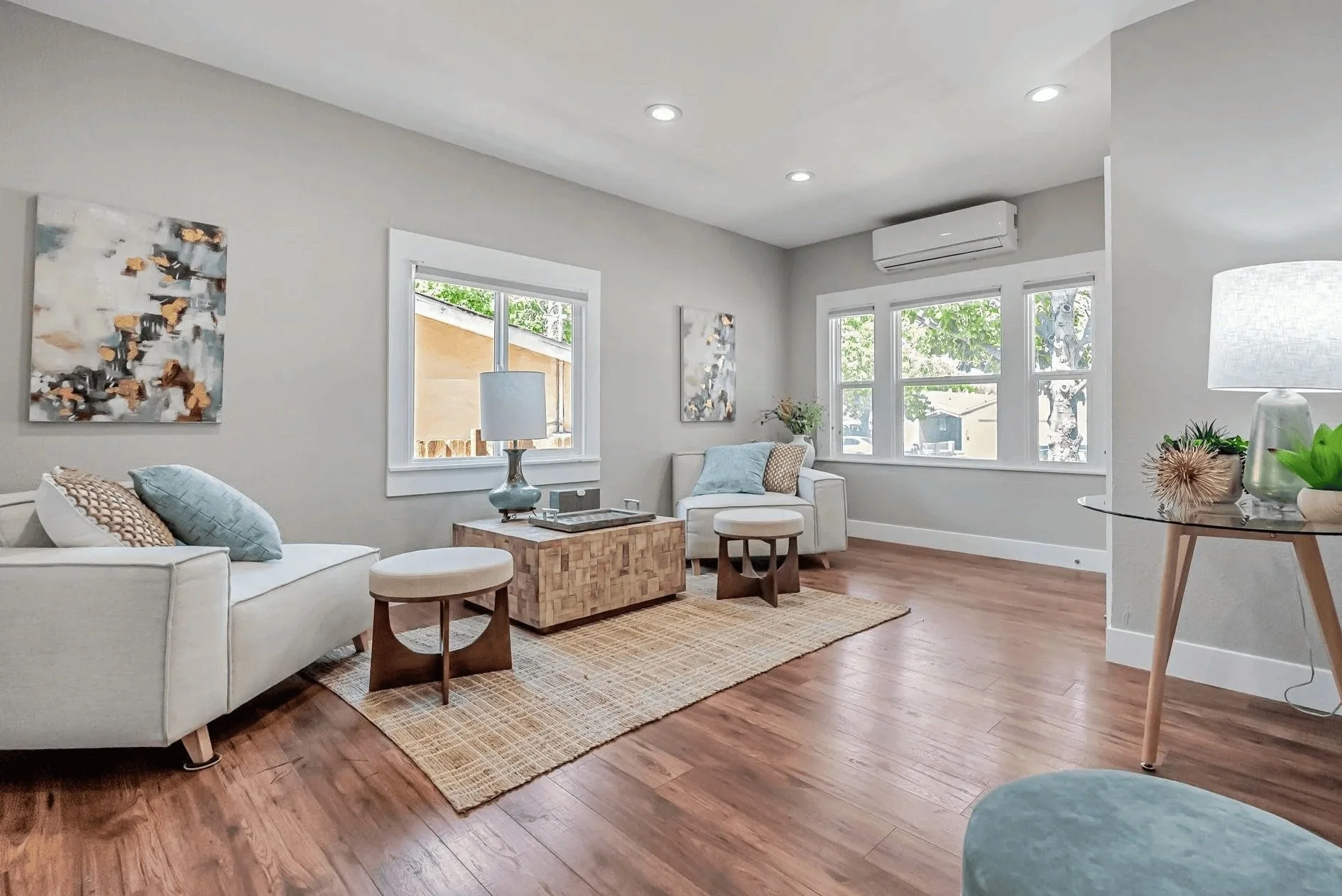 Bright living room with white sofa, armchair, wooden coffee table, textured rug, framed abstract art, lamps, window views of trees and neighboring houses, and hardwood floors.