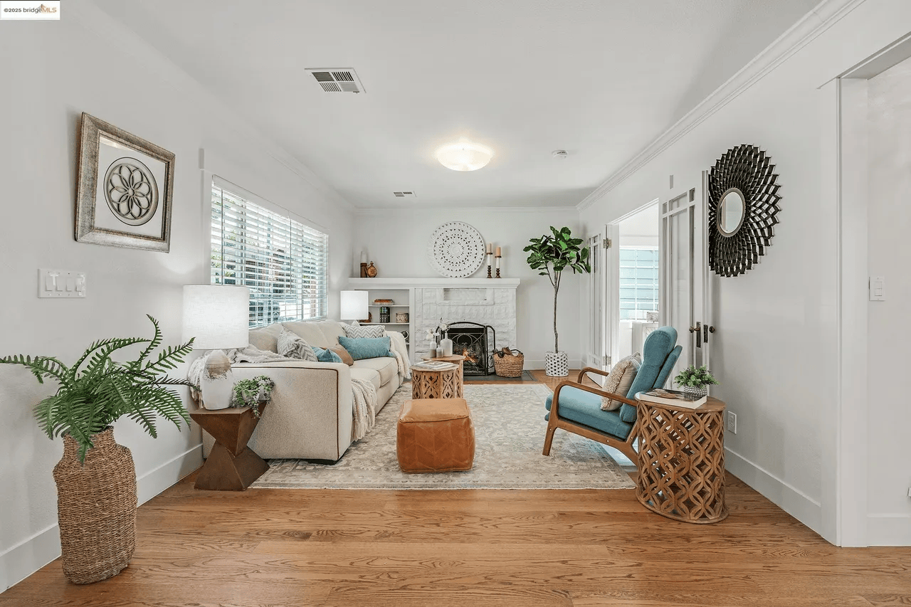 Bright living room with white walls, hardwood floors, beige sofa with blue and beige pillows, teal armchair, woven side tables, potted plants, fireplace, decorative wall mirrors, artwork, window blinds, and a cozy rug.