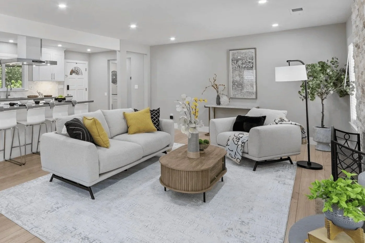 Modern living room with white sofa and armchair, a wooden coffee table, decorative pillows, plants, and artwork on the wall.
