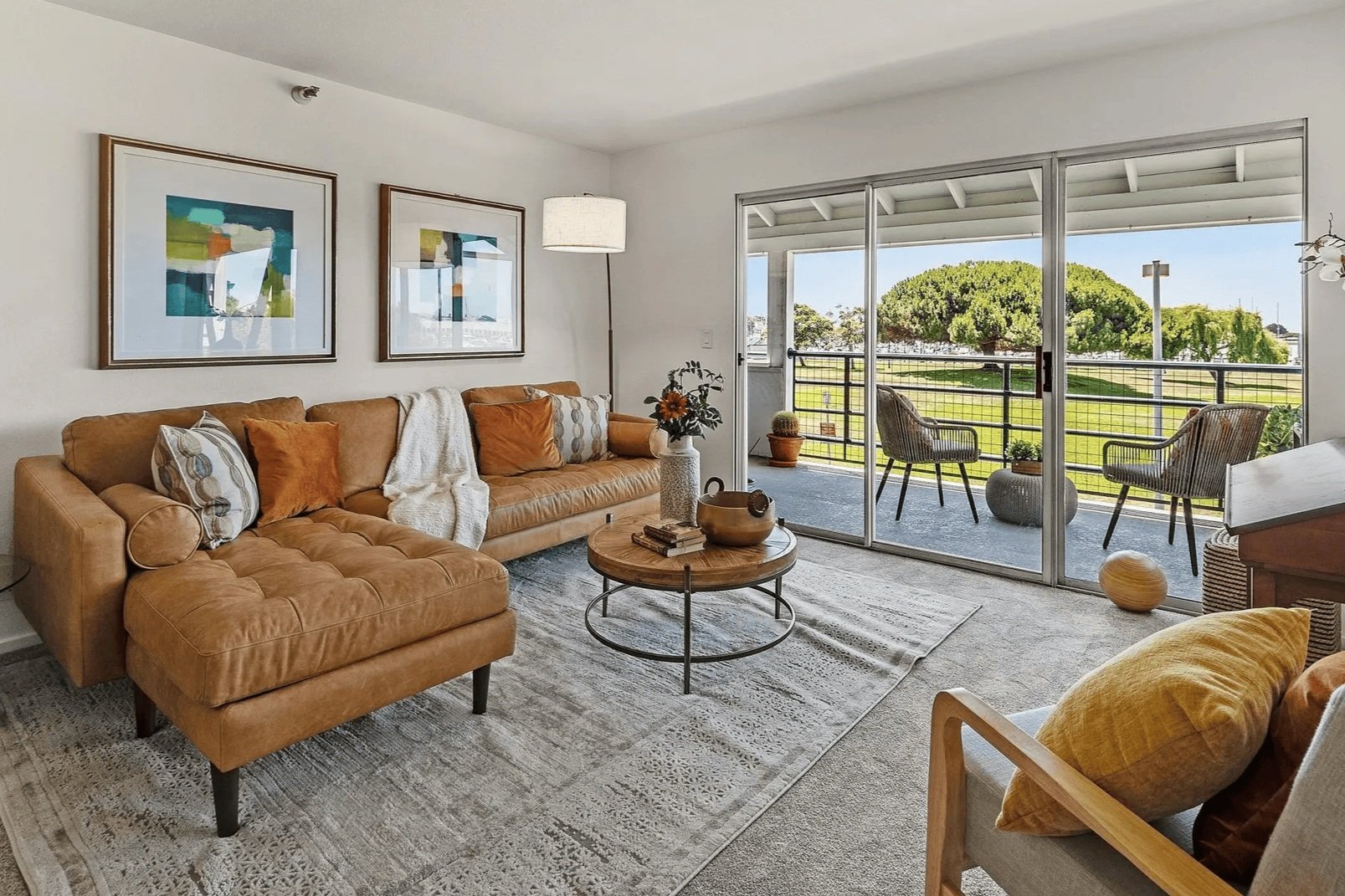 Living room with a tan leather sofa, decorative pillows, a white throw, a round wooden coffee table with decor, and a large window providing a view of a balcony with chairs and a green outdoor landscape.