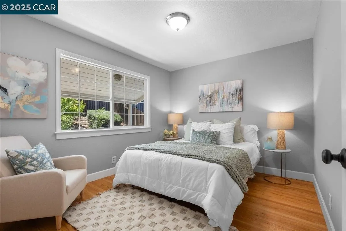 A cozy bedroom with a window showing greenery outside. The room features a white bed with multiple pillows, a beige armchair with a decorative pillow, two bedside lamps, and abstract artwork on the walls. The floor is wooden with a cream-colored rug.