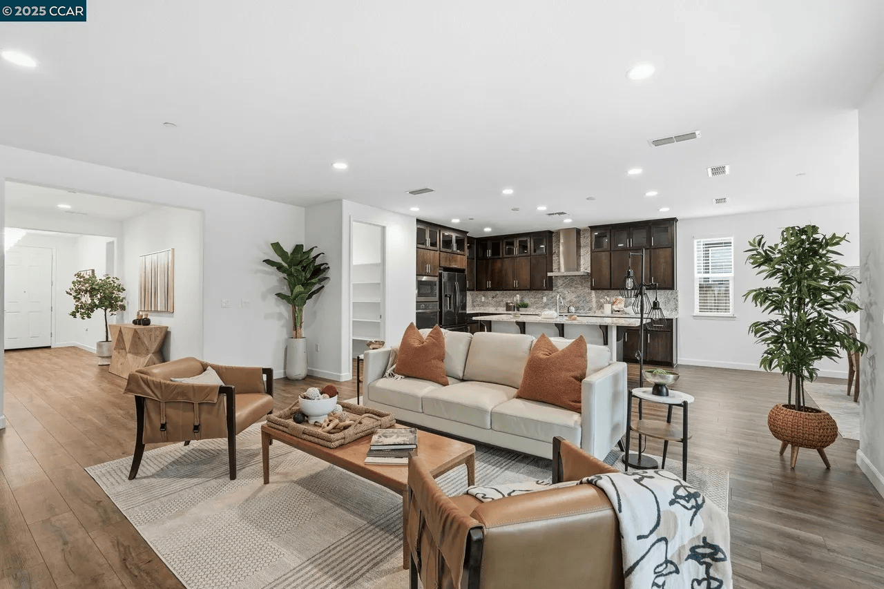 Open-concept living room with white sofa, tan armchairs, wooden coffee table, and kitchen with dark cabinets, granite countertops, and modern appliances. Potted plants and decorative pillows add style.