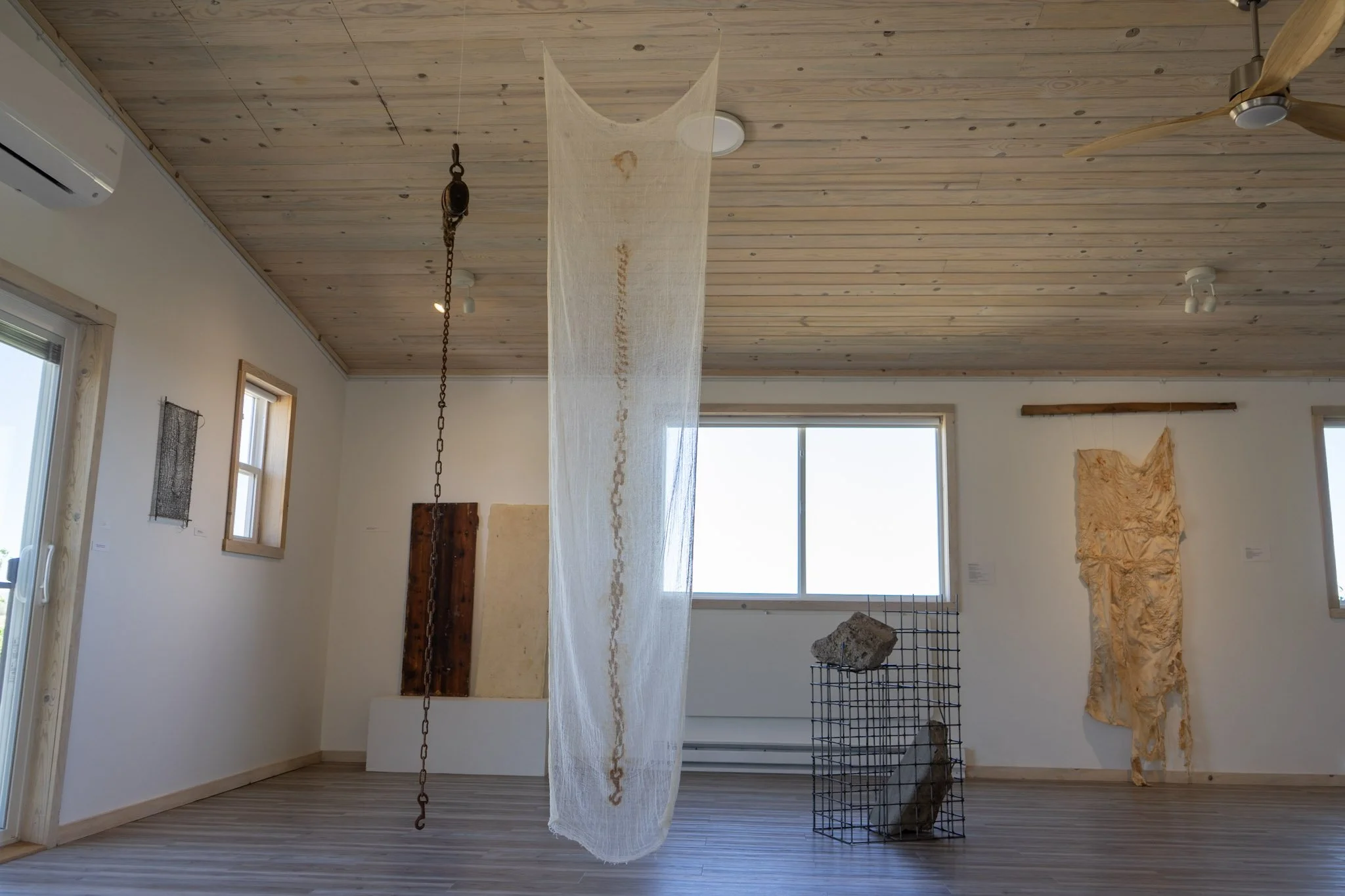 Interior view of an art gallery with wooden ceiling and floor, featuring hanging textile art, sculptures on wire frames, and art on the wall near windows.