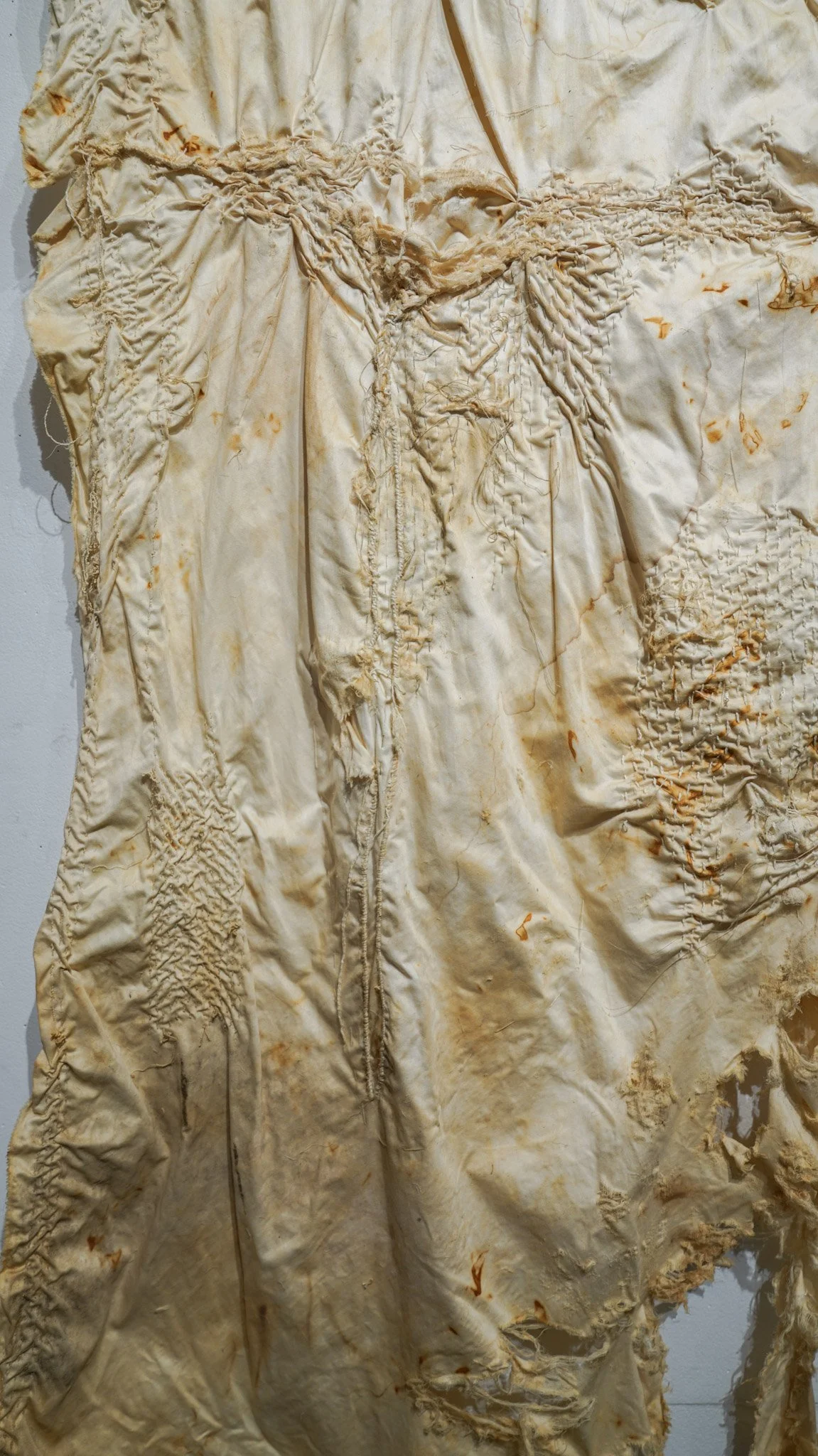 Tattered, aged, beige-colored fabric with stained, frayed, and ripped areas hanging against a plain white wall.