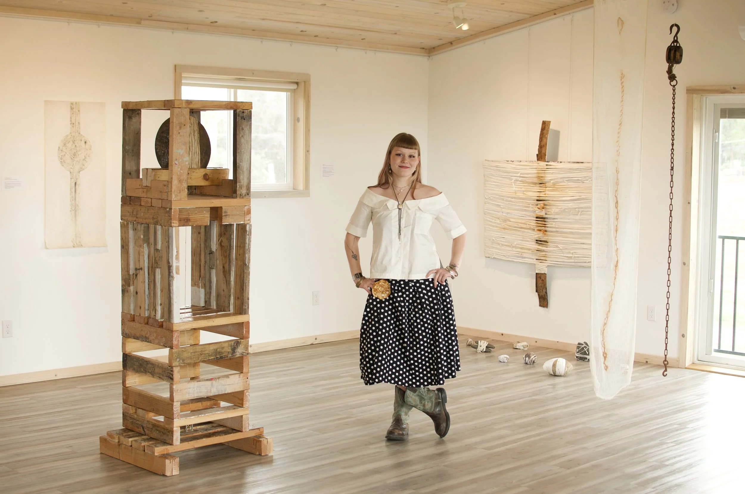 A woman with light brown hair, wearing a white off-shoulder top and polka dot skirt, stands in an art gallery. The gallery features wooden artwork and hanging sculptures, with white walls and wooden flooring.