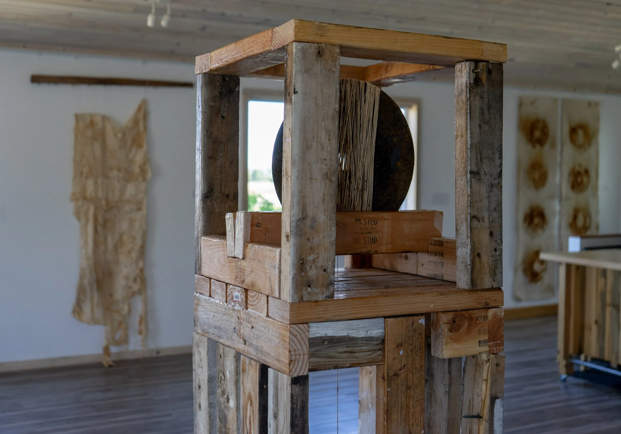 A wooden art sculpture resembling a small house or building frame inside a room with wooden floors and walls, and decorative wall hangings in the background.