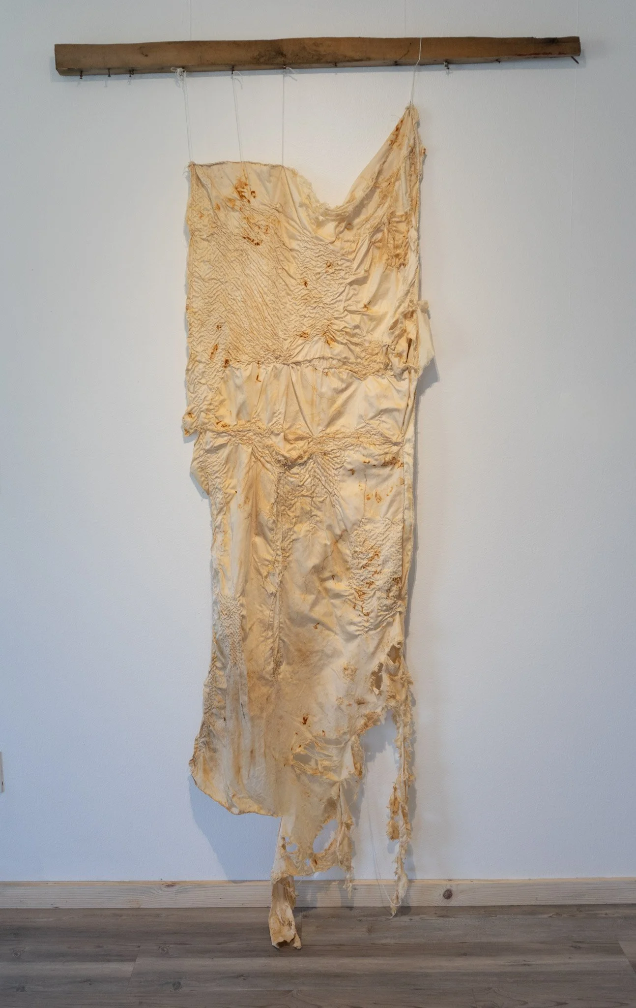 An art installation features a long, tattered, cream-colored fabric with rust-colored stains hanging from a wooden plank mounted on a white wall. The fabric appears crumpled and aged with irregular edges.