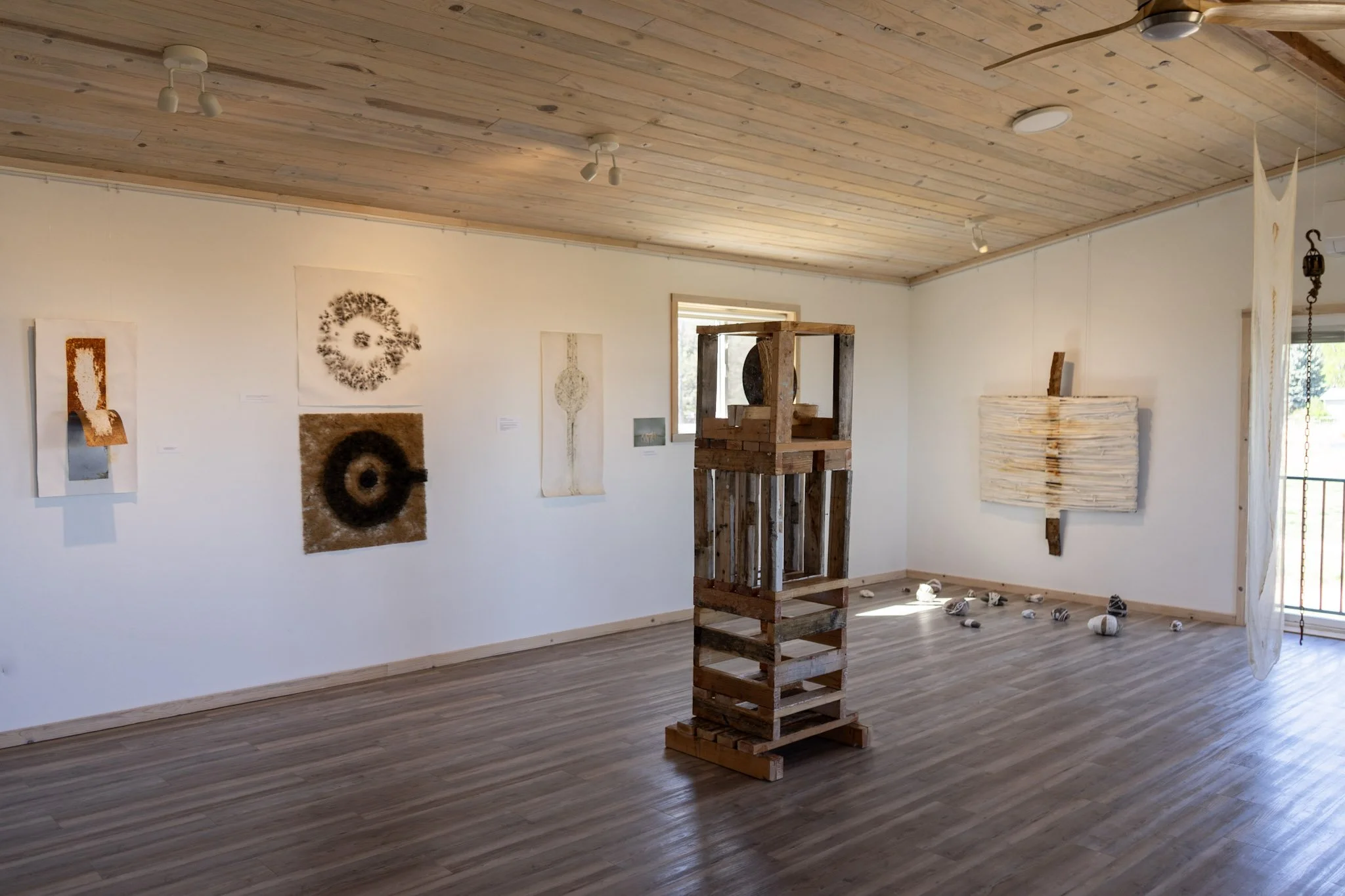An art gallery with wooden ceiling and white walls displaying various abstract art pieces, including circular and organic shapes. There are scattered objects on the floor and a wooden sculpture in the center.