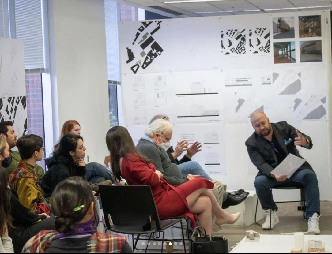A diverse group of people attending a presentation or discussion in a room with architectural plans and photos on the wall.