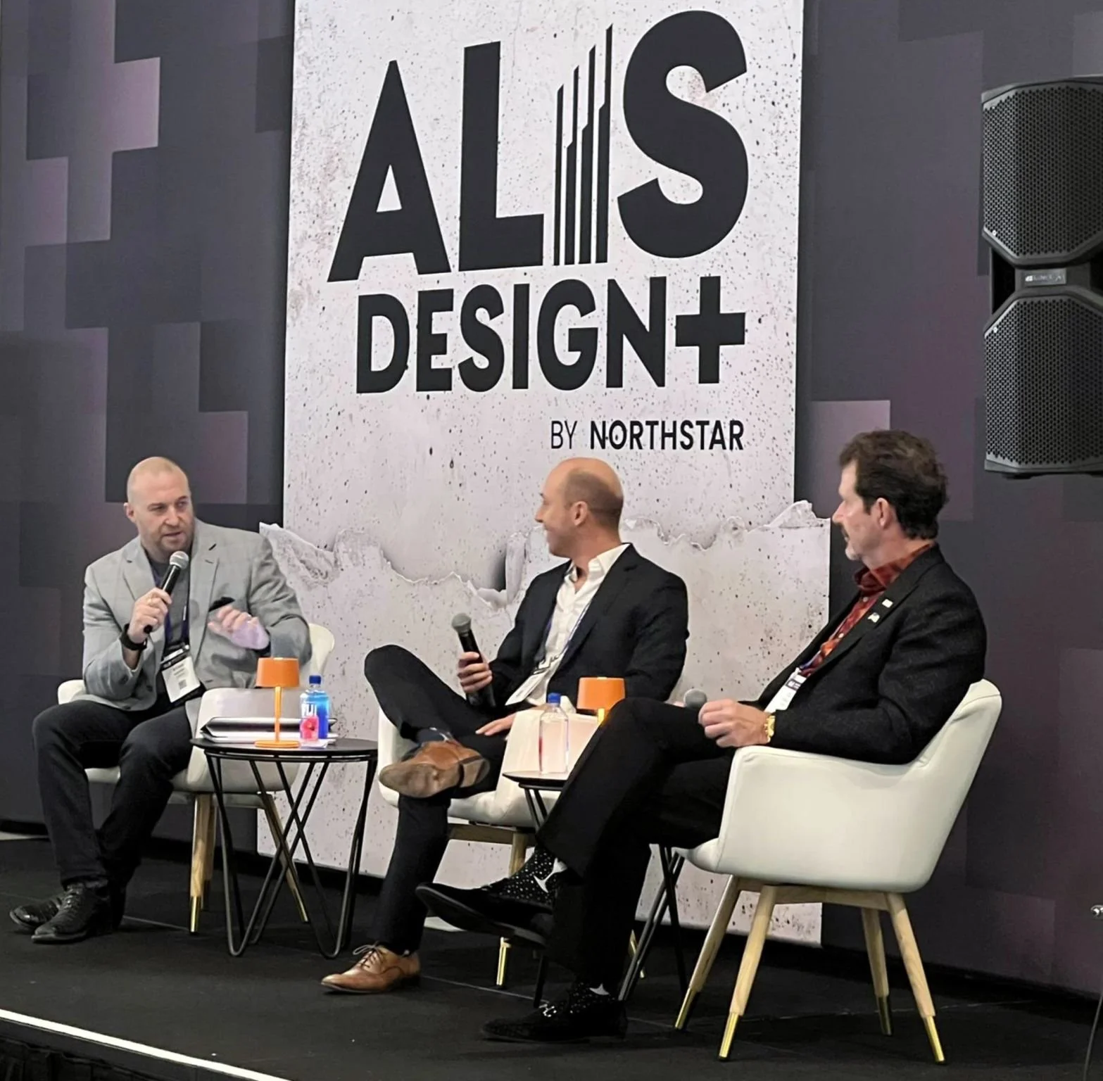 Three men in suits sitting on stage in front of a large sign that says 'ALIS DESIGN by NORTHSTAR,' engaged in a panel discussion, with microphones and small tables with drinks in front of them.