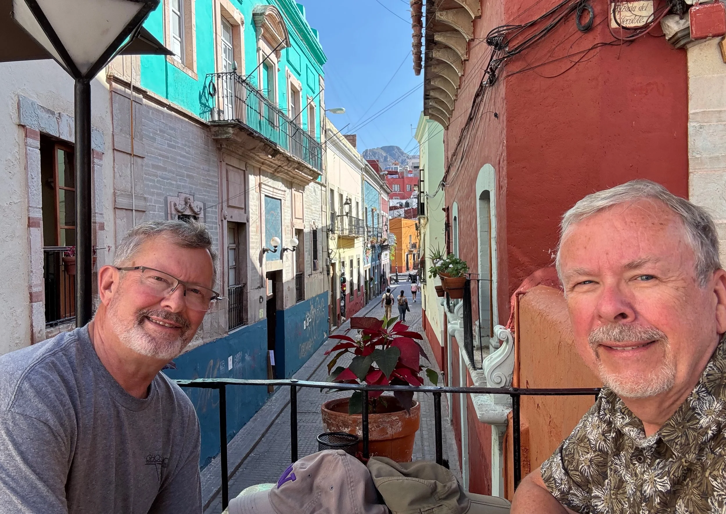 Two smiling men sitting on a balcony overlooking a colorful street with small buildings, some with balconies and plants, in a hilly cityscape under a clear blue sky.