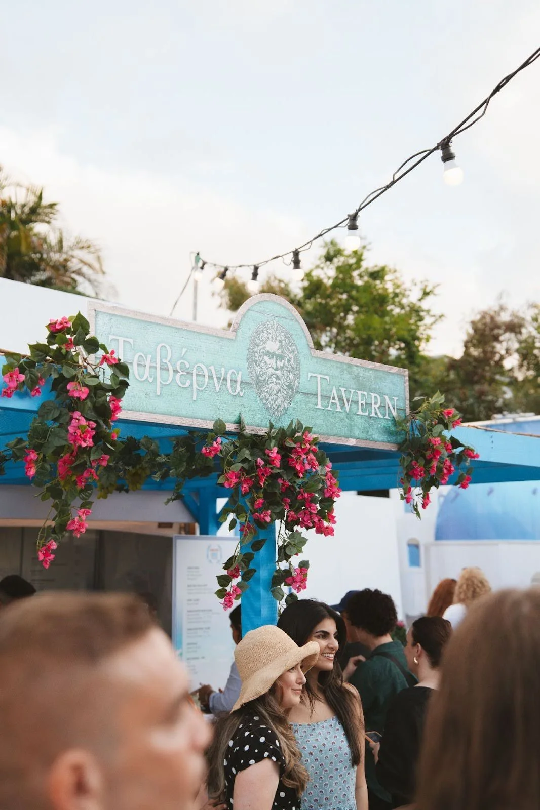 People gather outside a blue Greek-style tavern decorated with pink flowers and greenery, under string lights, during daytime.
