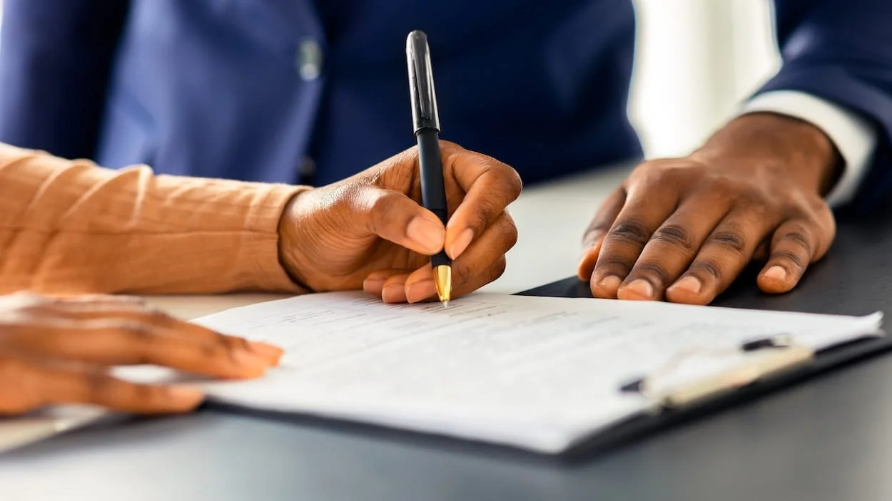 A person is signing a document with a black pen held in their right hand, while another person is resting their left hand on the document.